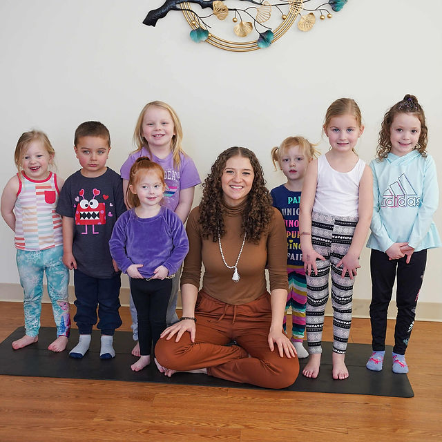 Group of eight children and one adult woman sitting and standing on a yoga mat indoors, smiling, with a decorative wall hanging above.