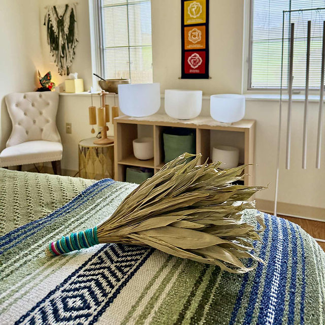 Dried plant on a bed with a colorful blanket, in a room with a white chair, wooden shelf, and window.