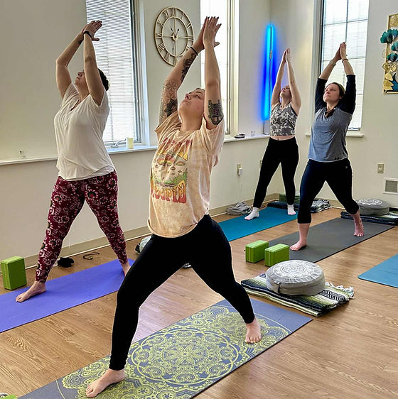 Four people practicing yoga in a room with windows, mats, and props, stretching with arms raised overhead.