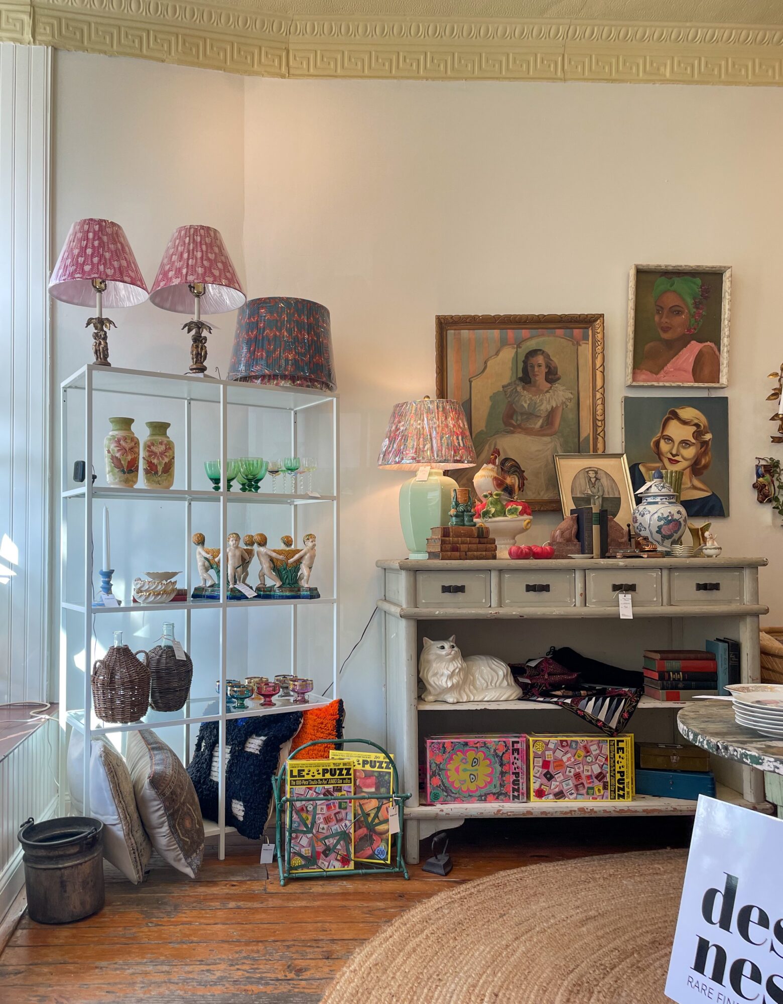 Living room corner with shelves, framed artwork, lamps, and decorative items, including a cat figurine and colorful boxes.