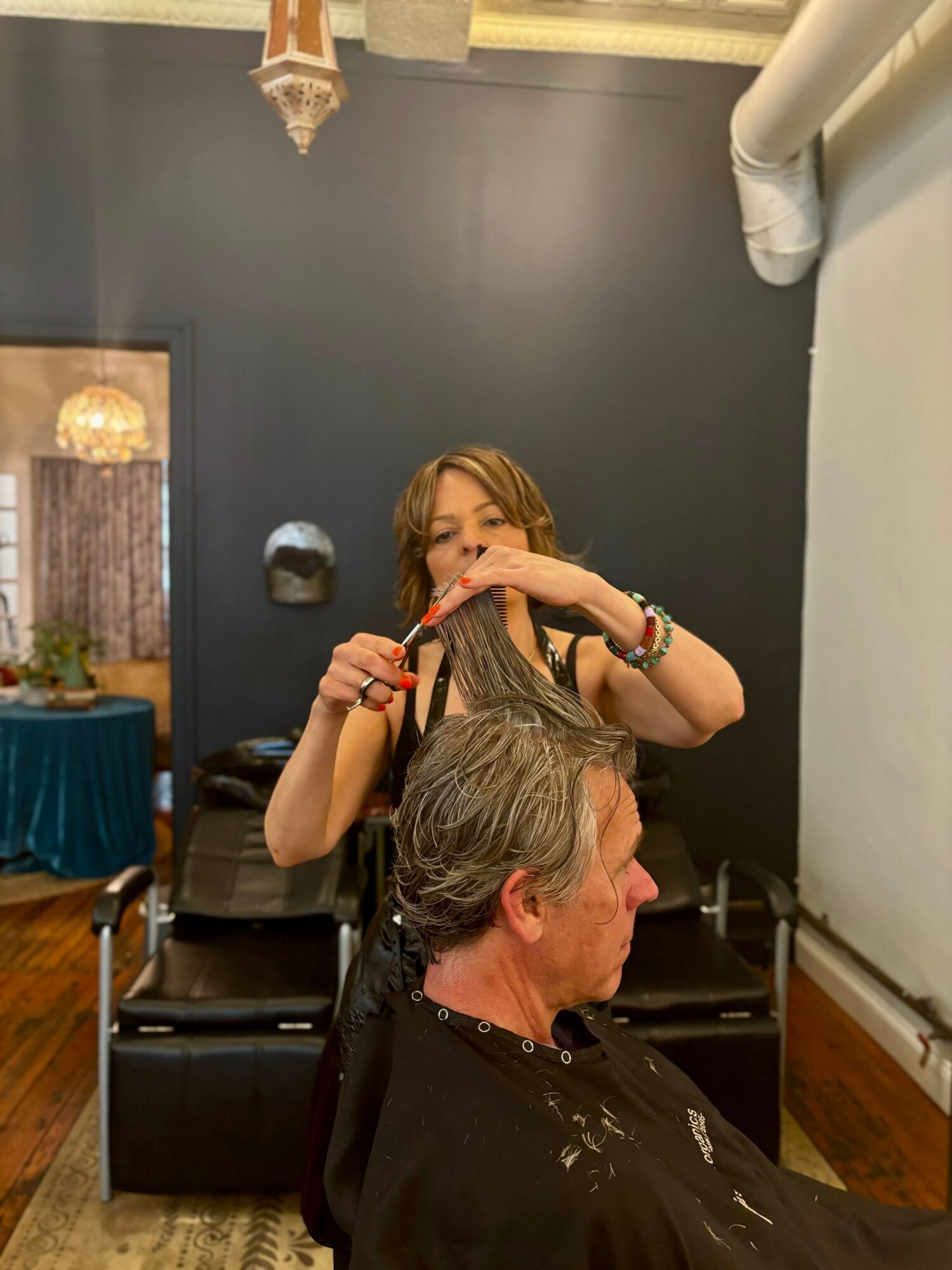 Woman with long hair and bracelets trims a man's hair in a salon, mirror reflects the scene, dark wall background.