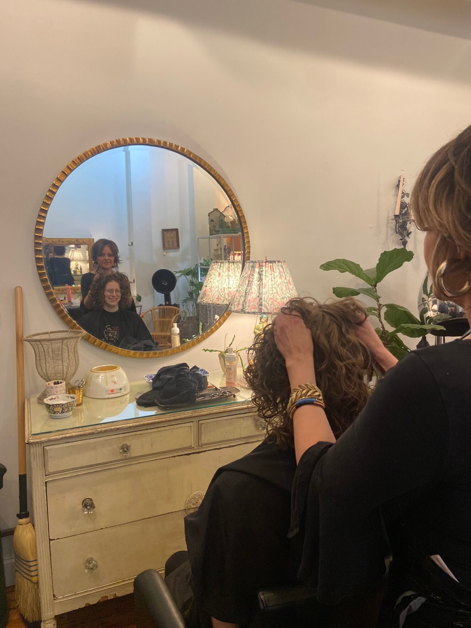 Person getting a haircut in a salon, with a mirror reflecting two people and salon decor, including a lamp and plants.