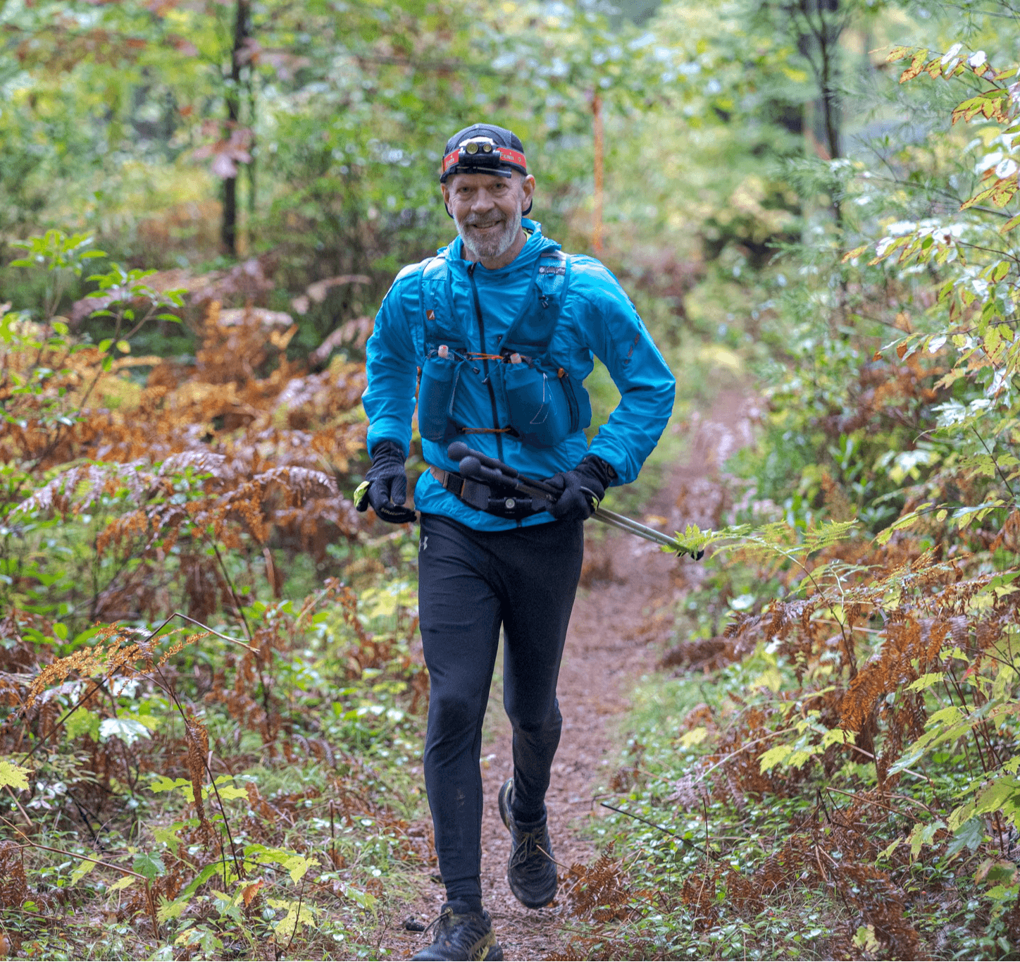 Man running on a forest trail, wearing a blue jacket, black pants, and a headband, surrounded by green and brown foliage.