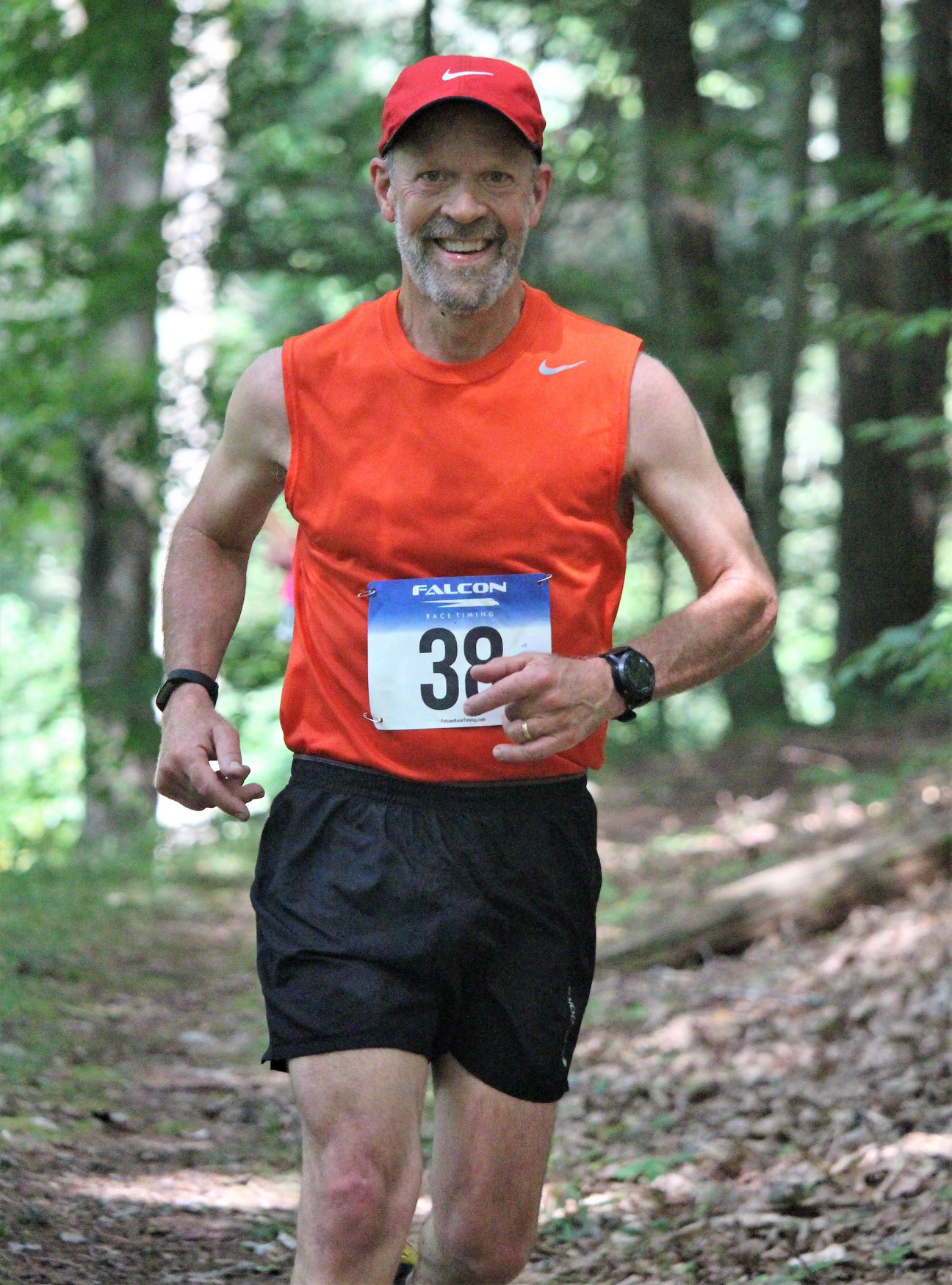 Man running in a forest, wearing a red cap, orange sleeveless shirt, black shorts, and a race bib number 39.