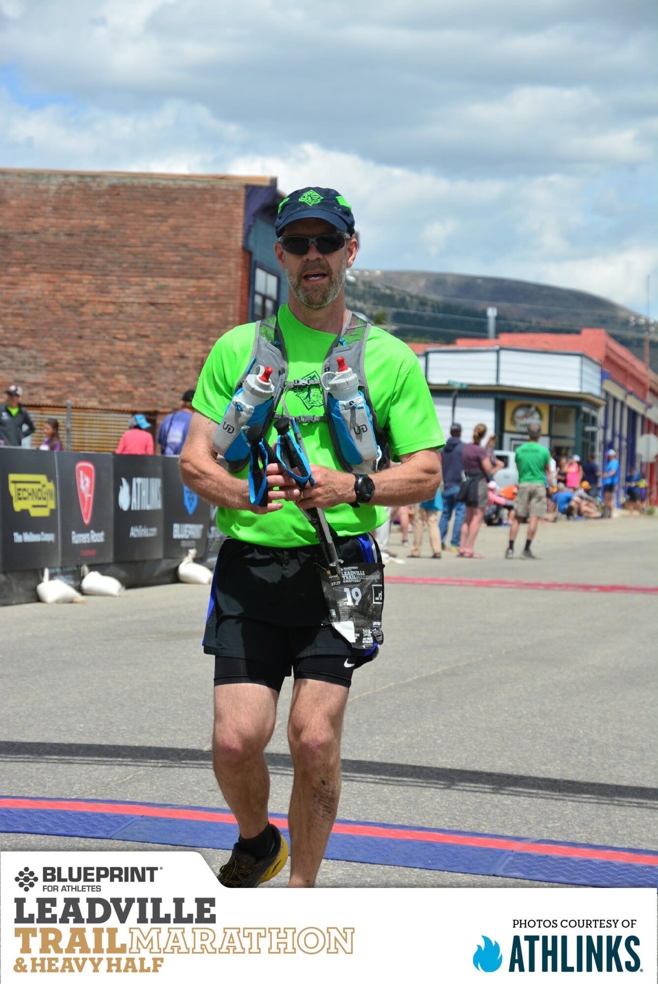 Male runner in bright green shirt and black shorts holding water bottles, walking outdoors with people and buildings in background.