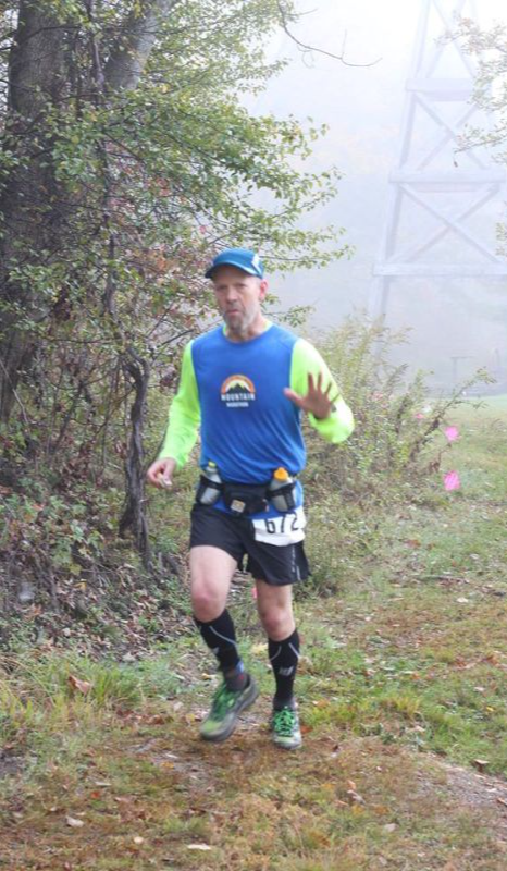 Runner in blue shirt and black shorts running outdoors on a trail with trees and fog in the background.