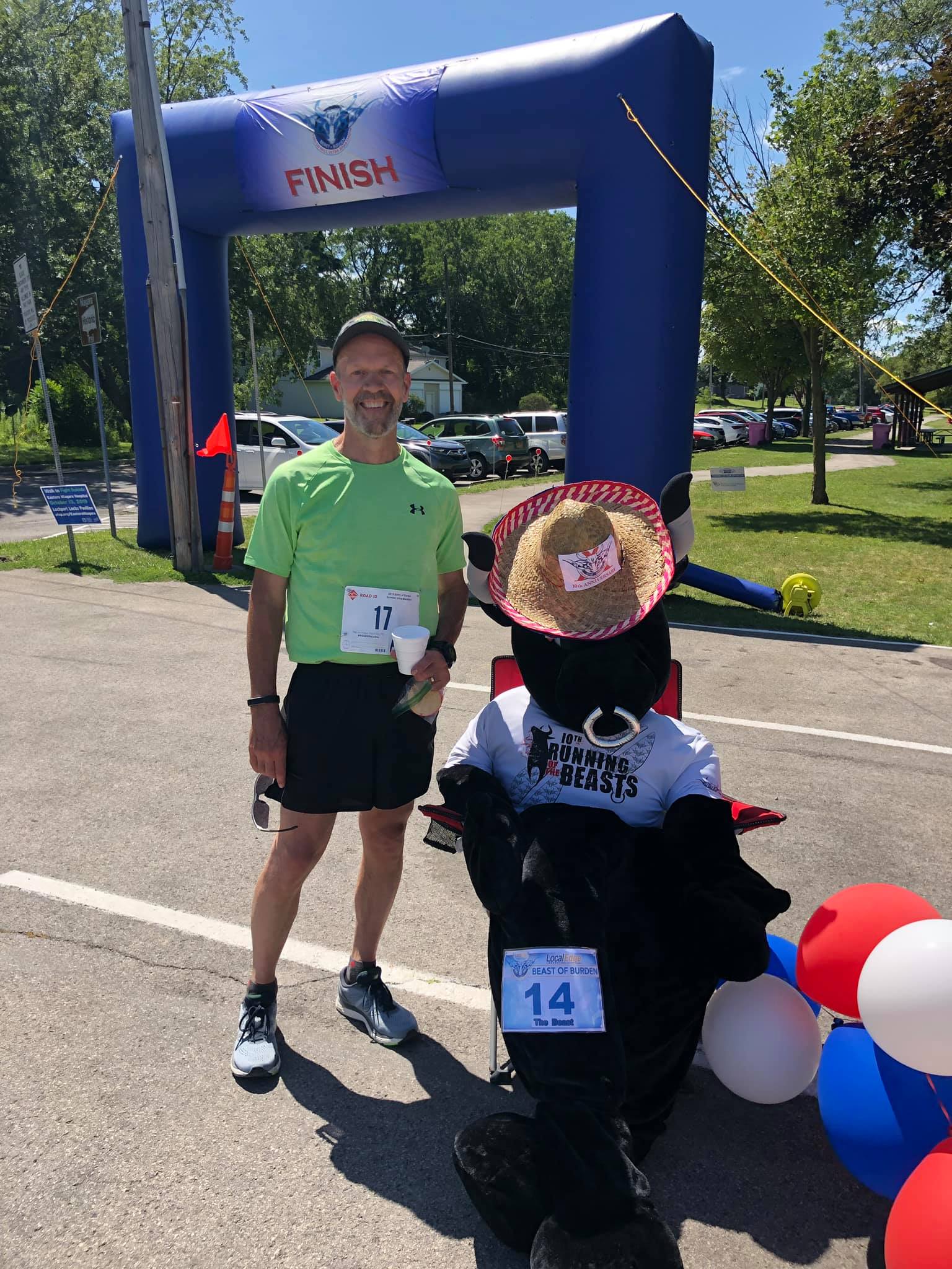 Man in green shirt and black shorts standing near finish line arch, person in costume with hat and balloons nearby, outdoor setting.