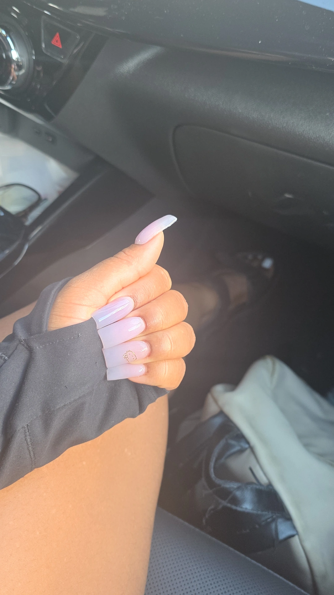 Person's hand with long nails giving a thumbs-up inside a car, dashboard and seat visible.