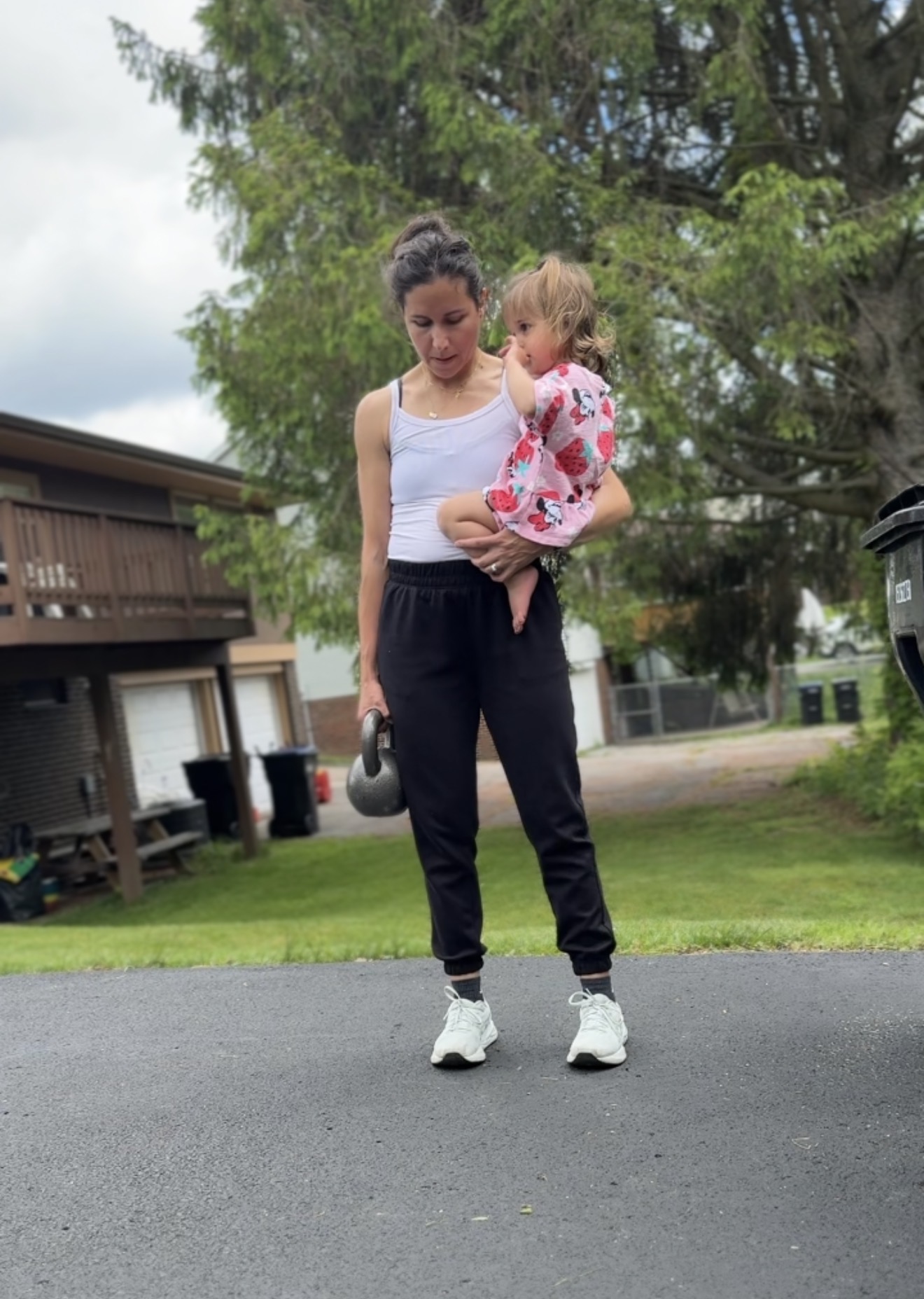 Woman holding a child outdoors on a street, with trees and houses in the background.