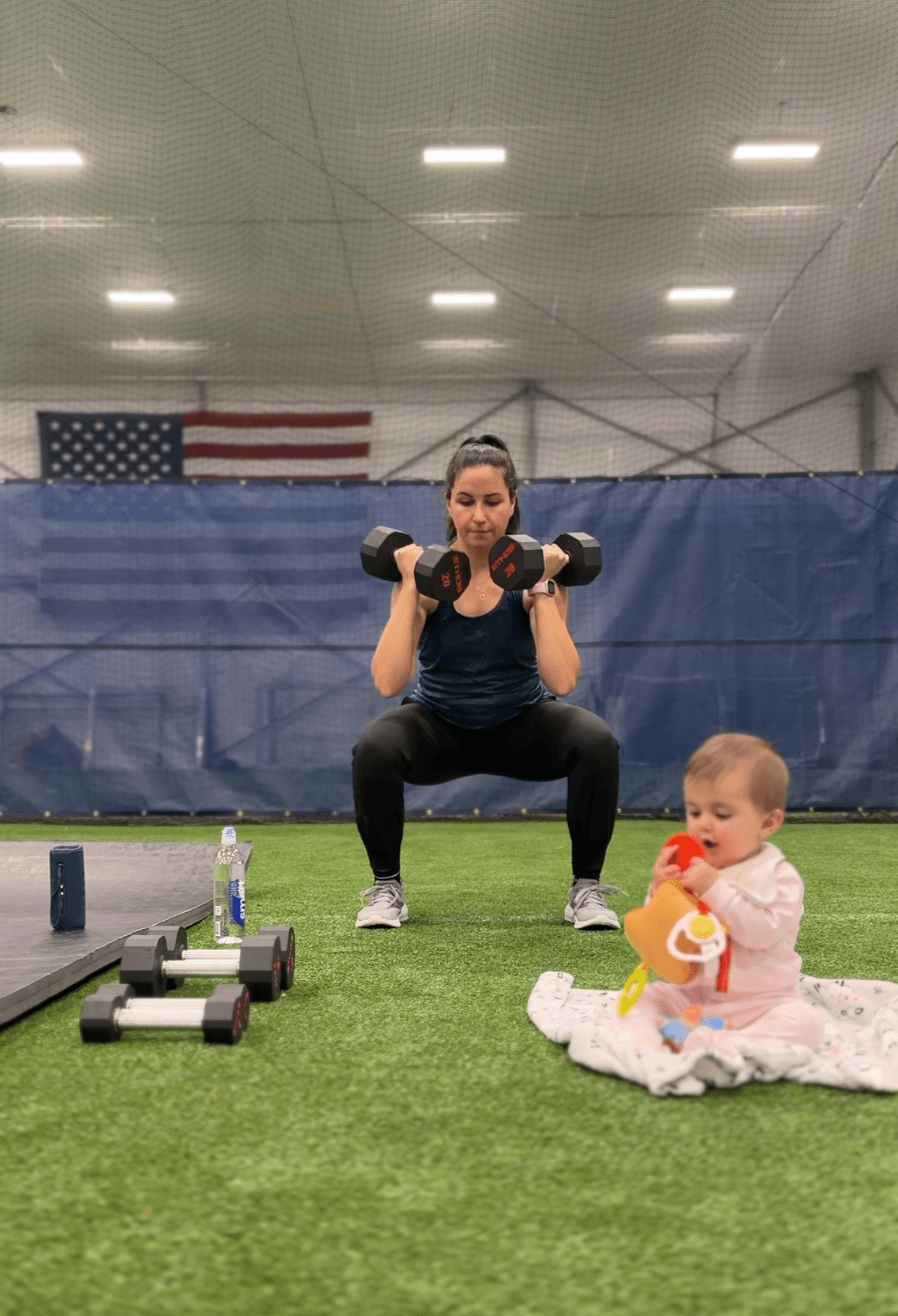 Woman squatting with dumbbells in an indoor gym, child sitting on the floor holding a toy, American flag in background.