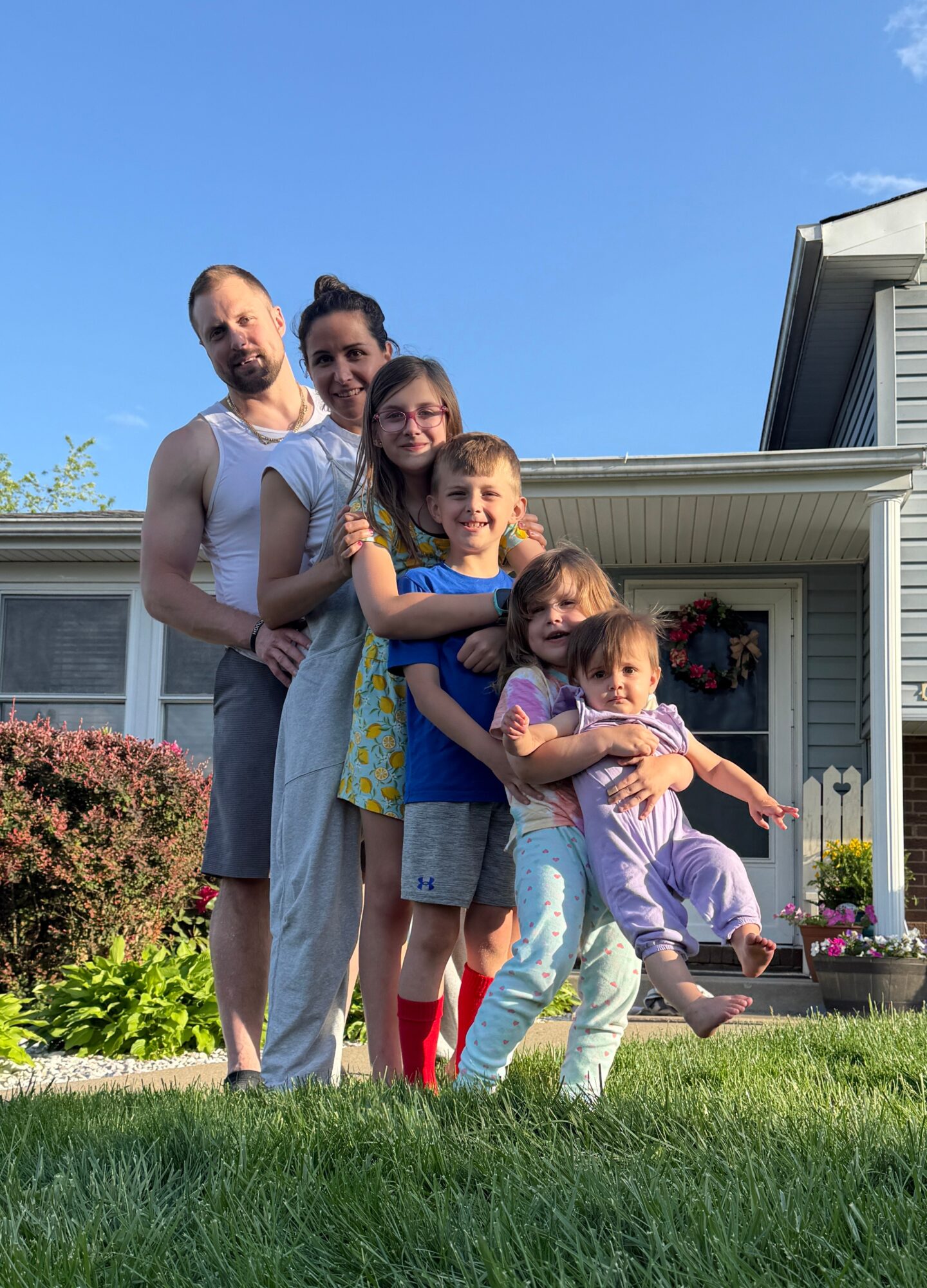 Group of six people, including children and adults, standing outside a house with a blue sky background.