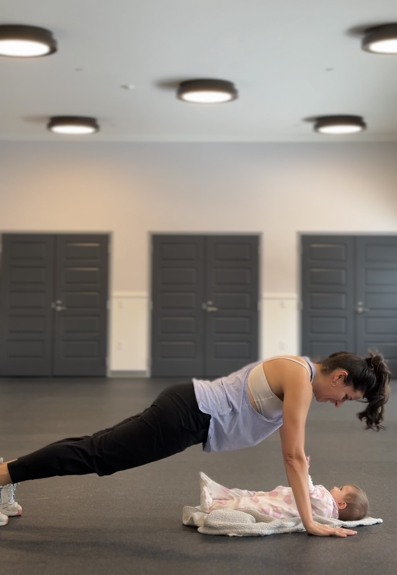 Person in a plank position on the floor with a baby, in a room with three closed doors and ceiling lights.