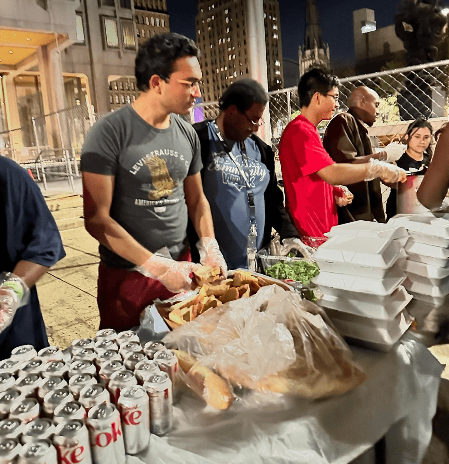People serving food outdoors at night, with city buildings in the background, stacks of food containers, and canned drinks on the table.