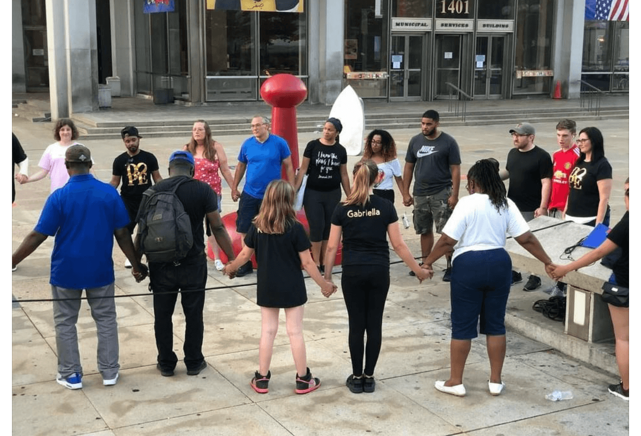 Group of people holding hands in a circle outside a building with glass windows and doors.