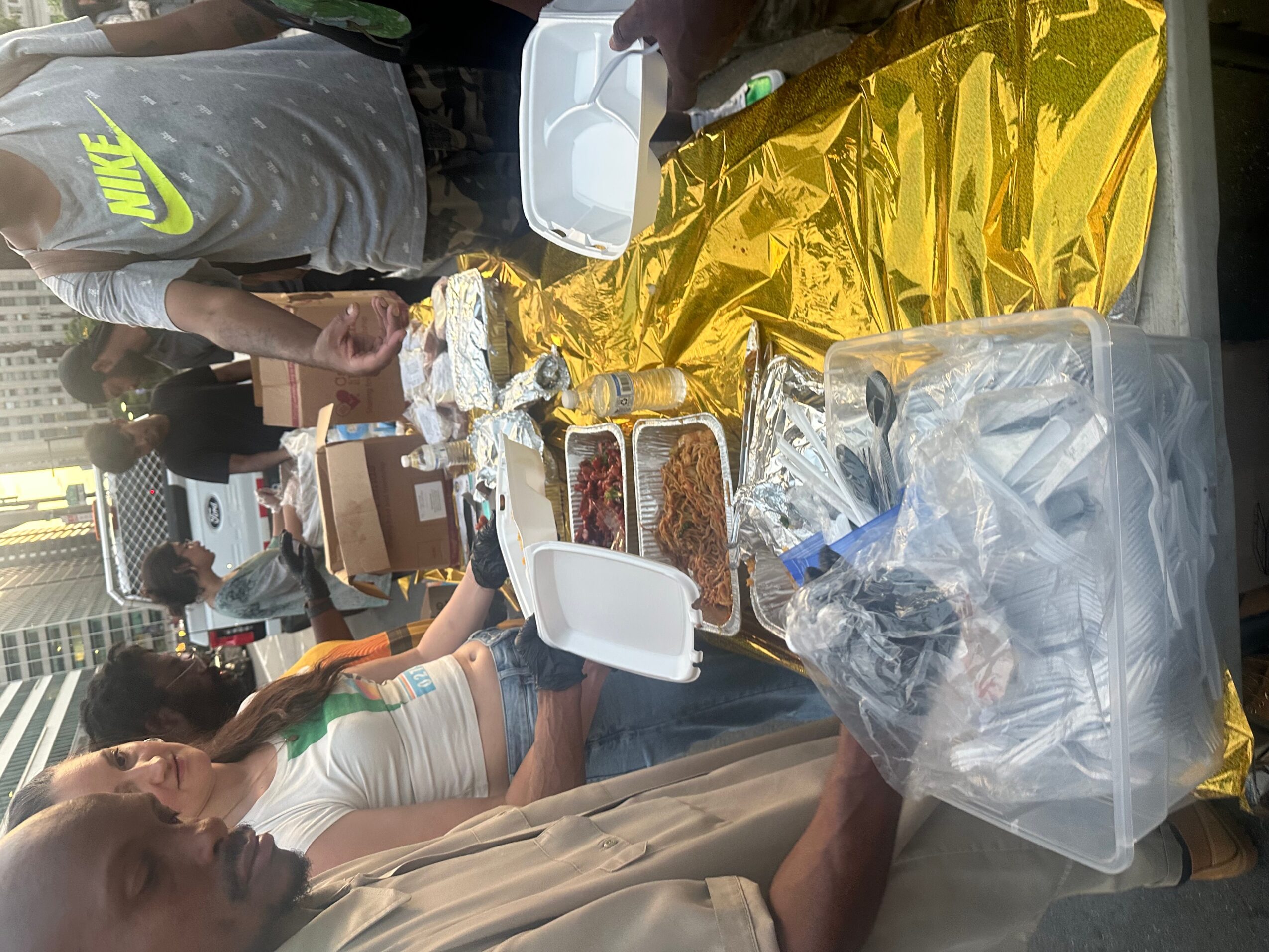 People gathered around a table with food, some sitting and some standing, outdoors during daytime.