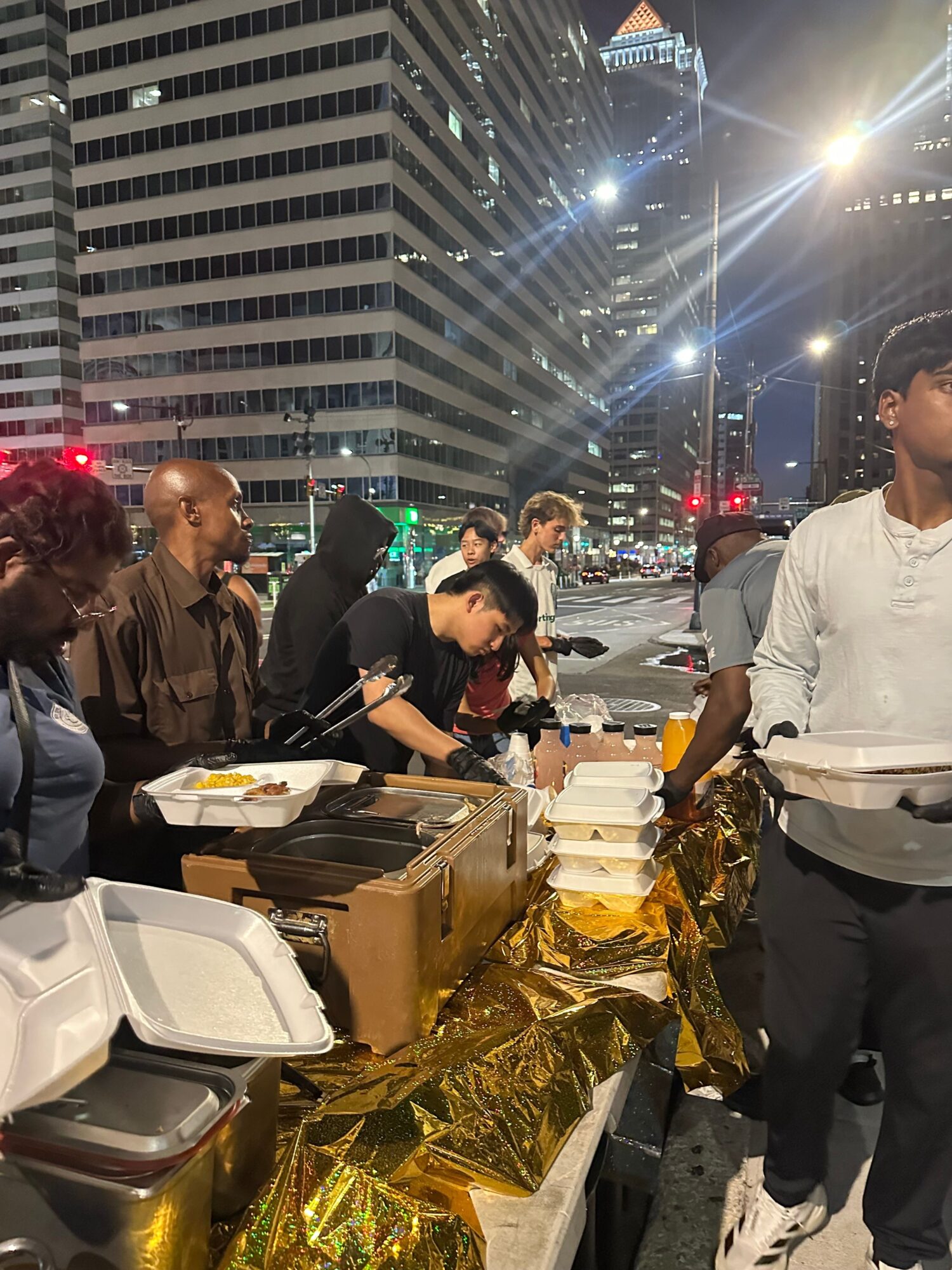 People gathered around a table with food and gifts in an urban nighttime setting with tall buildings and city lights.