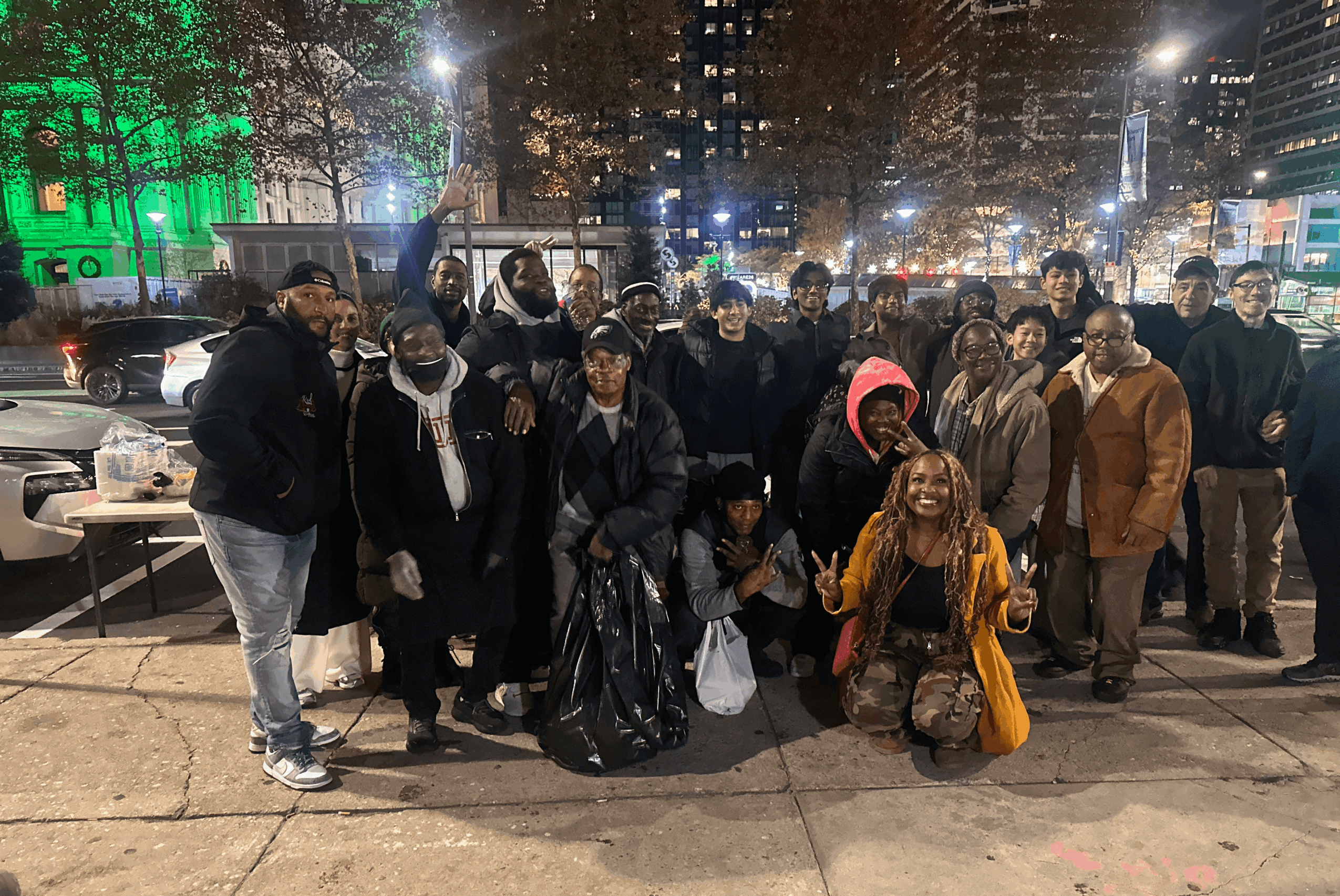 Group of people standing on a city sidewalk at night with buildings and lights in the background.