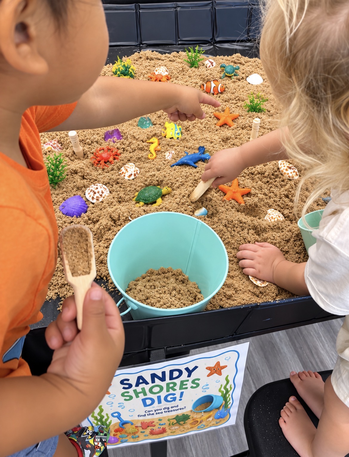 Two children play in a sandbox with toys, a blue bucket, and a sign reading 'Sandy Shores Dig!'.