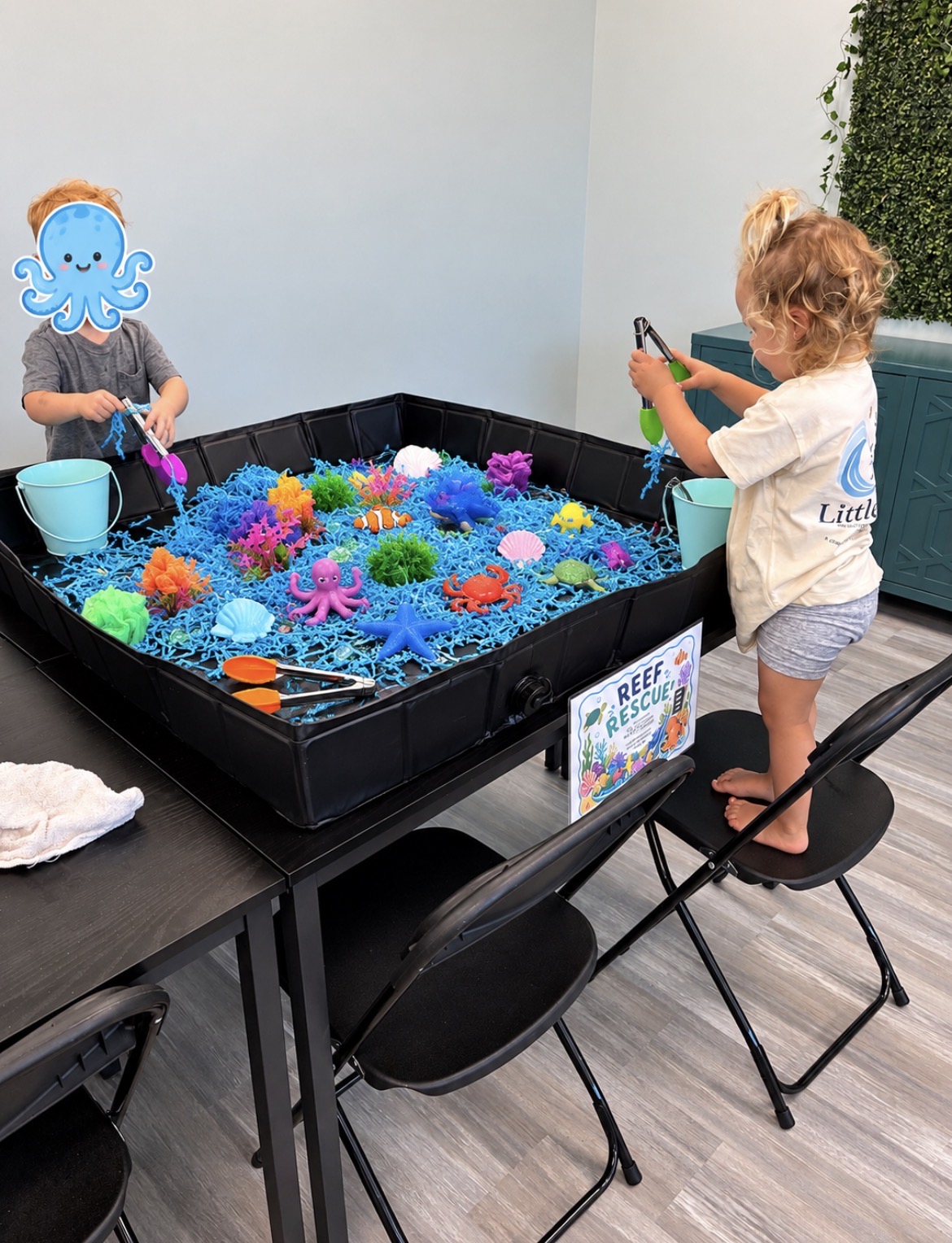 Two children playing with colorful plastic sea creatures in a small indoor water table, one with a cartoon octopus face sticker, on a table with chairs.