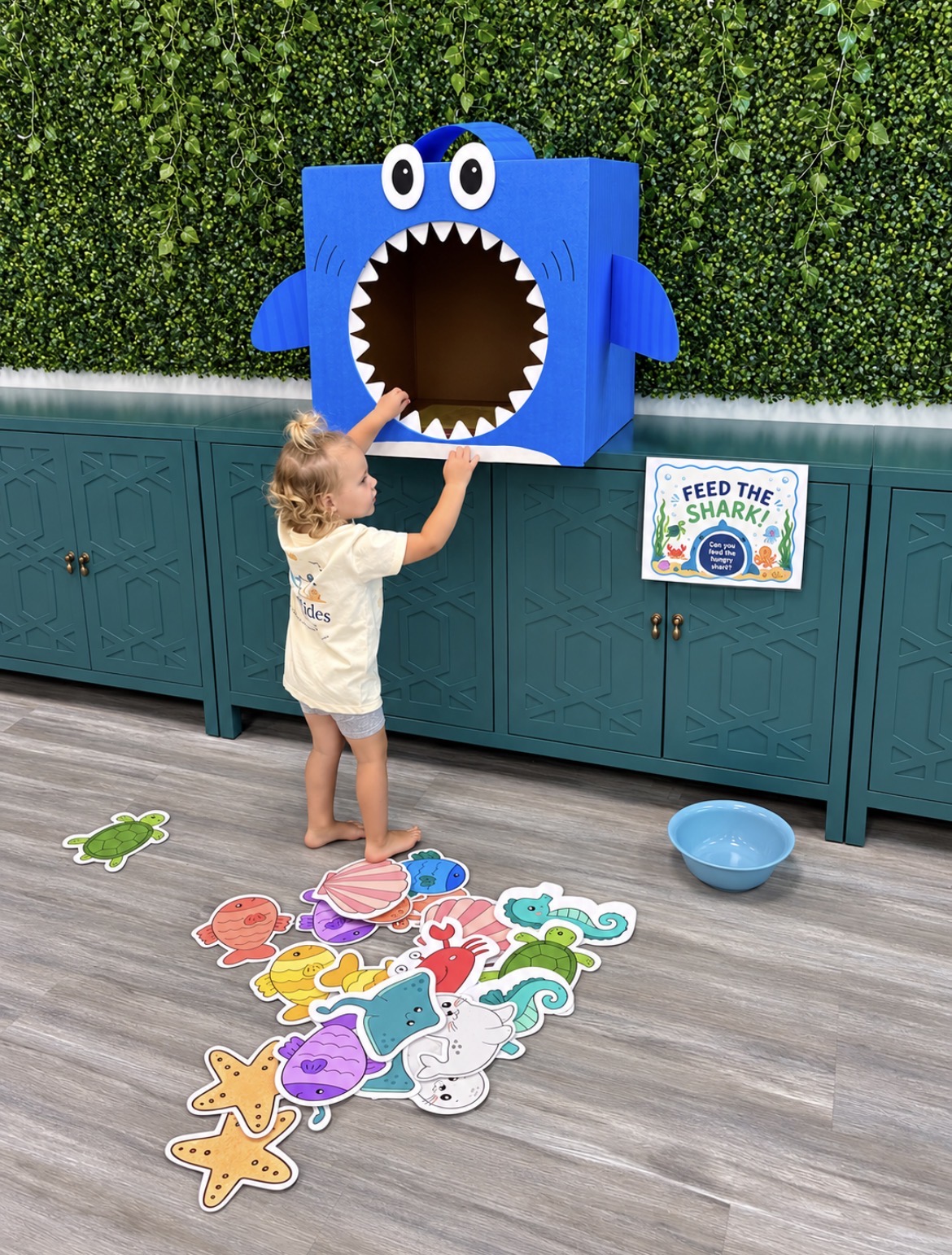 Child reaching into a blue shark-shaped box with a large open mouth, surrounded by colorful paper cutouts on the floor.