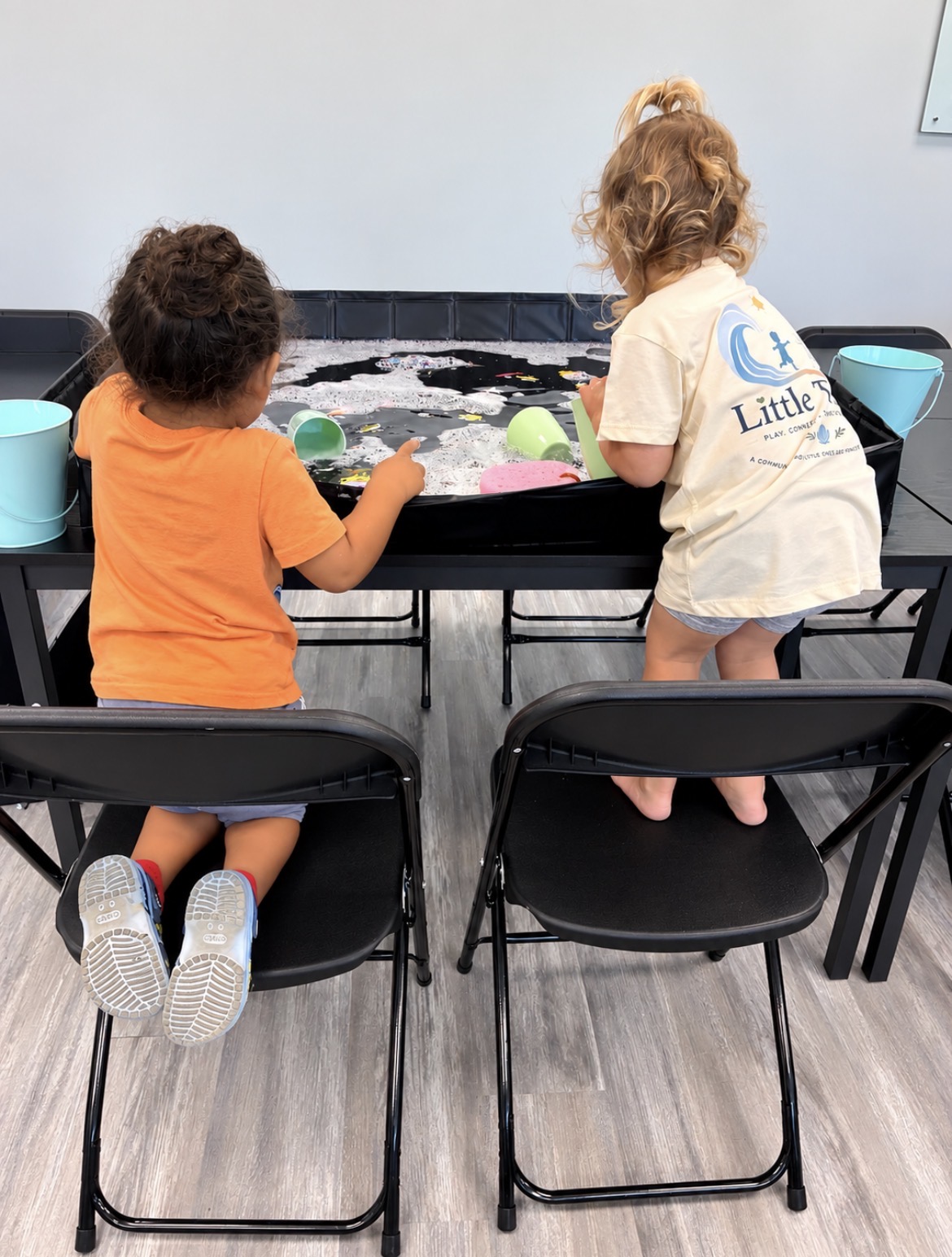 Two children, one with curly hair and one with a ponytail, play at a sensory table with foam and colorful objects.