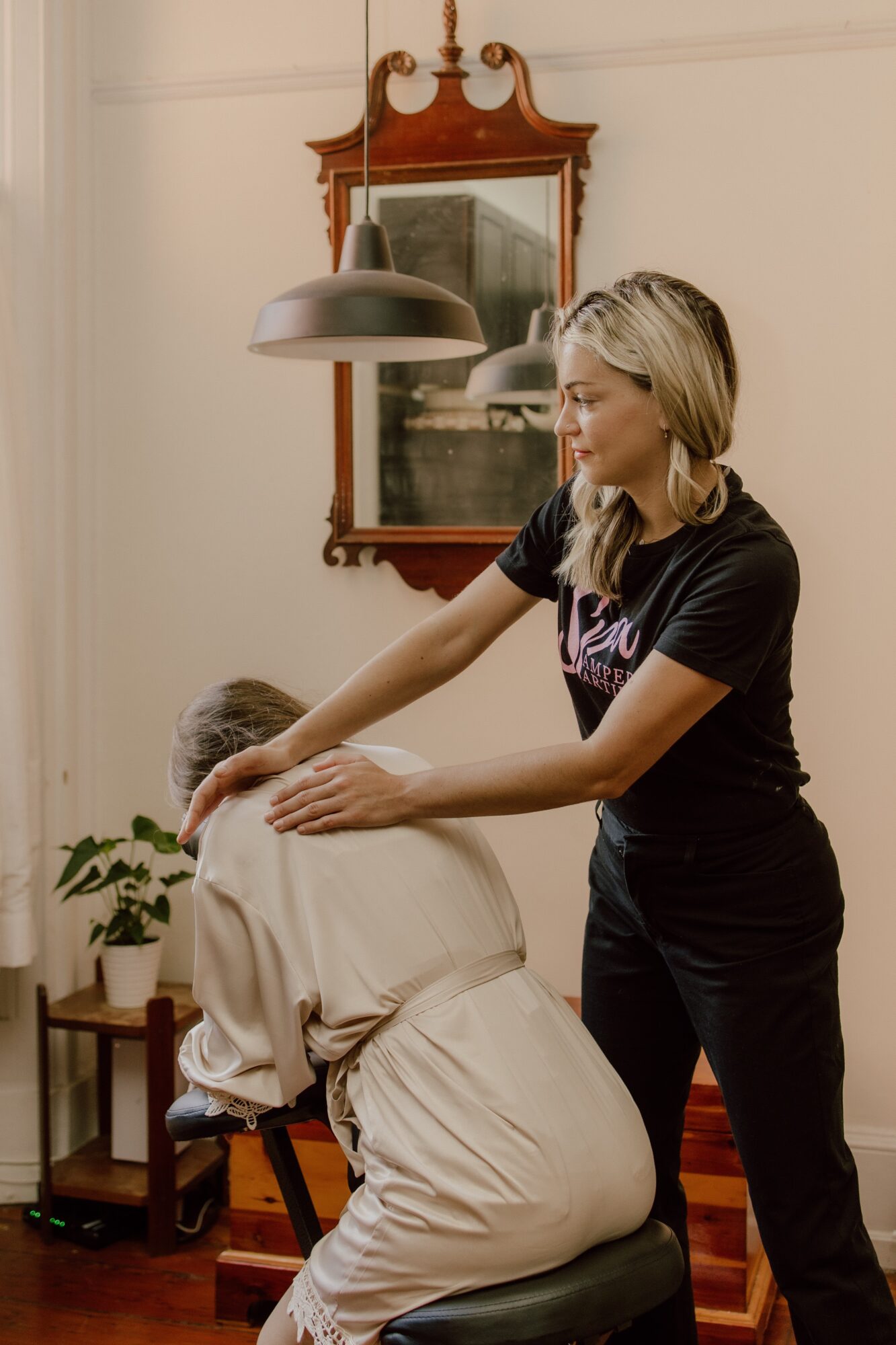 Woman giving a massage to a person seated in a chair in a room with a mirror and plant.