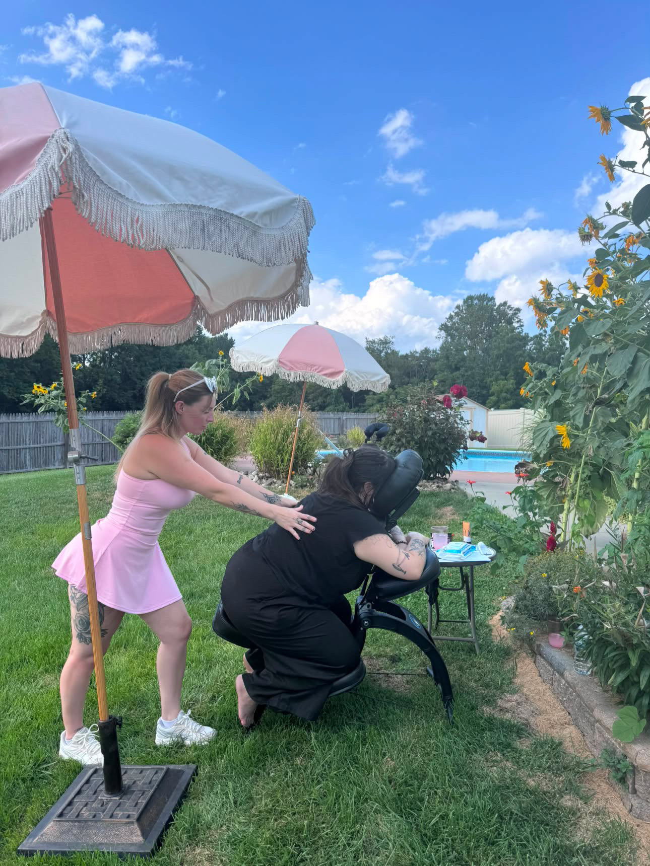 Two women outdoors near a garden, one in a pink dress and the other in black, with umbrellas and a pool in background.