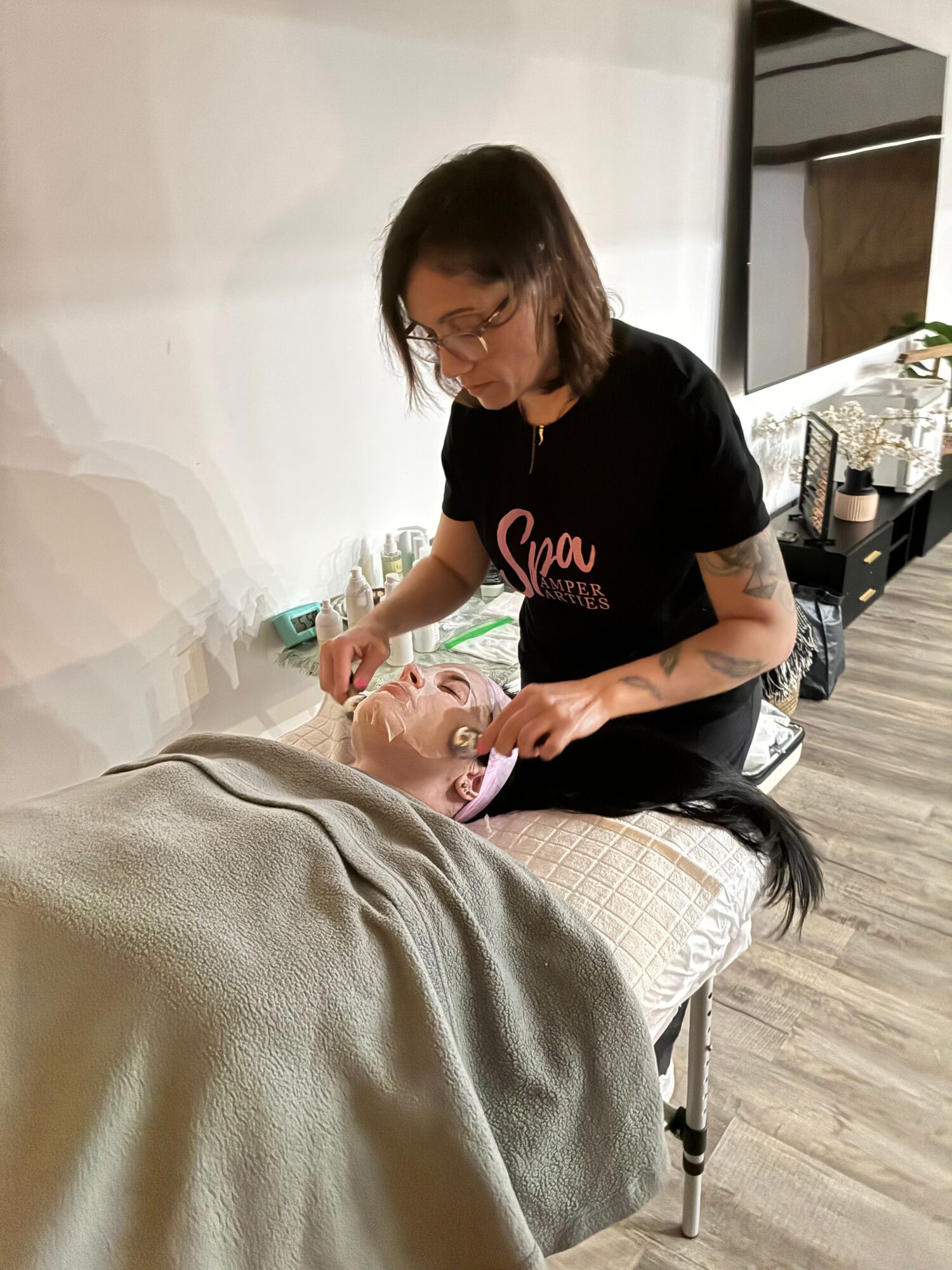 Person receiving a facial treatment lying on a bed, with a woman applying a mask or cream to their face in a spa setting.