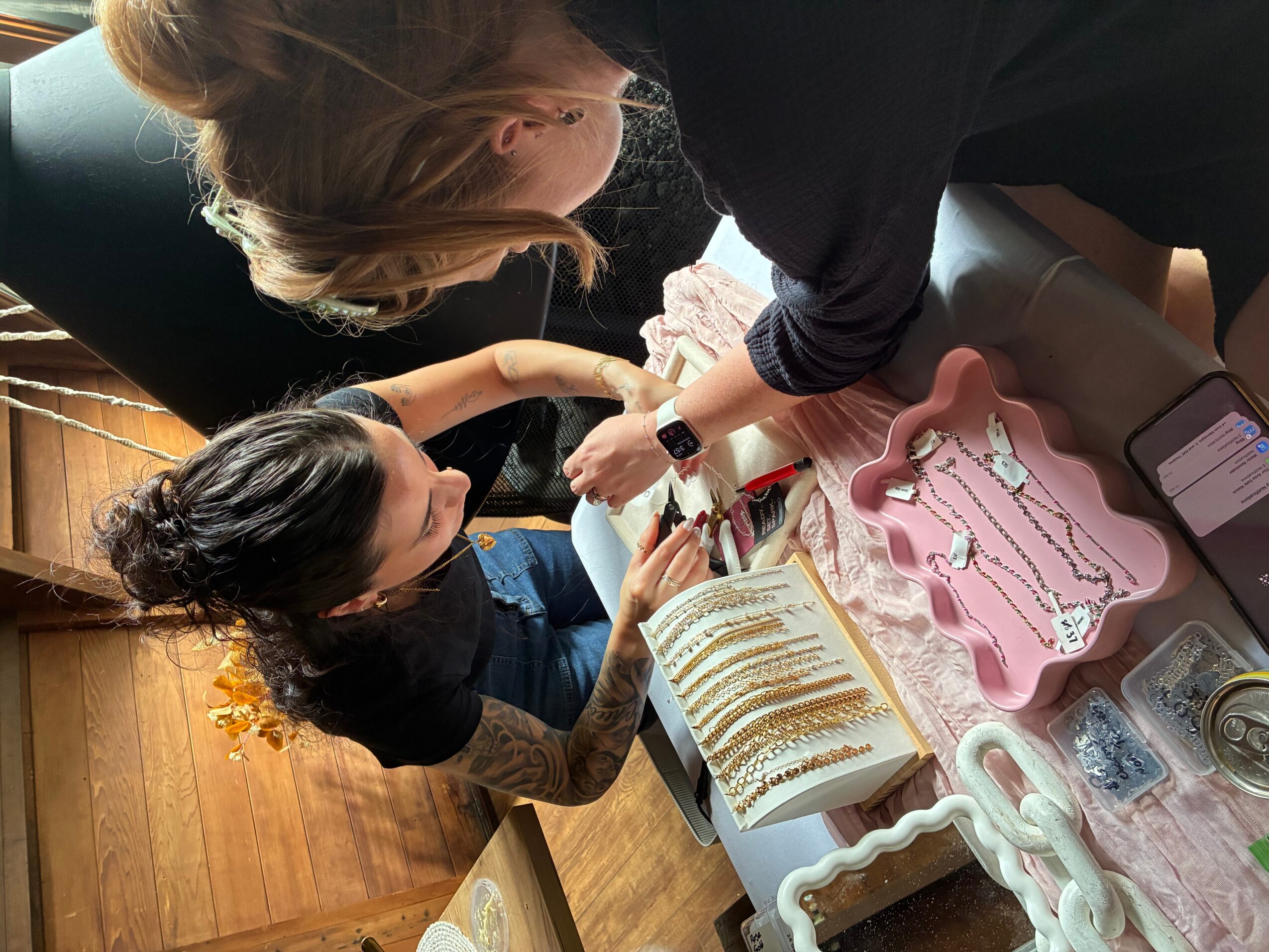 Two women are decorating a cake with gold accents, surrounded by baking tools and supplies on a pink tablecloth.