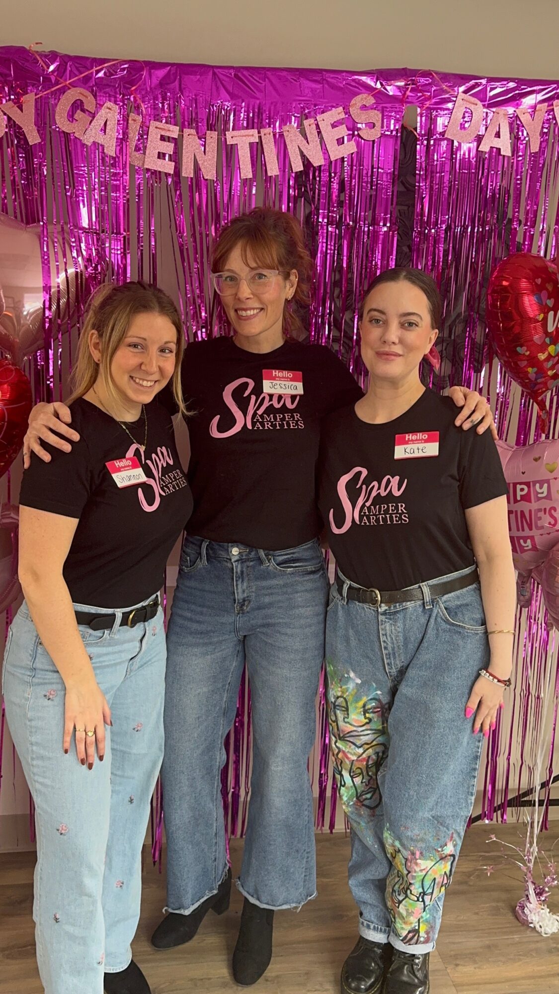 Three women standing together in front of pink and purple decorations, smiling, wearing black shirts with pink and white text, and jeans.
