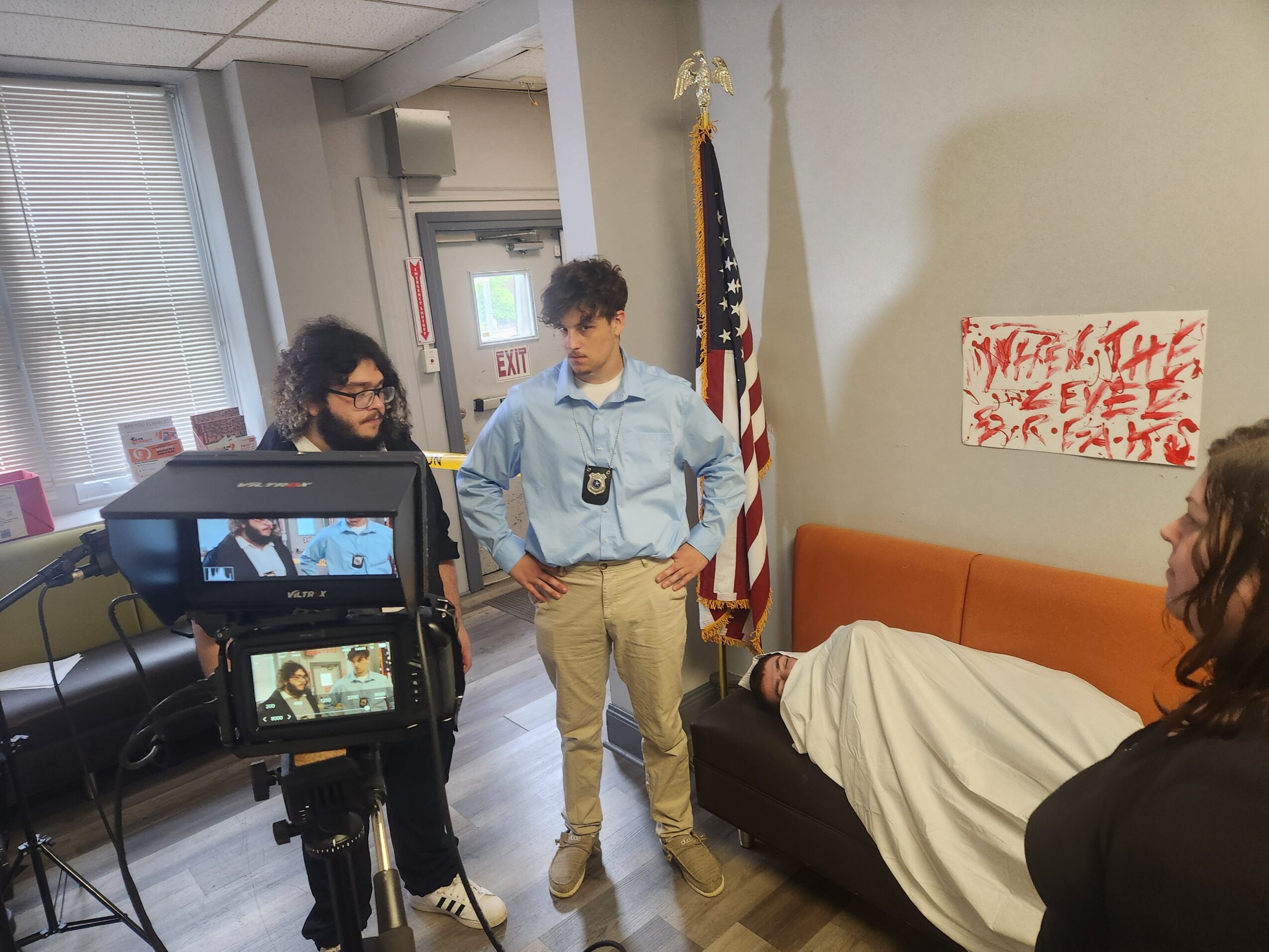 Person in security uniform standing next to a covered object on a couch, with two women and a camera recording the scene.