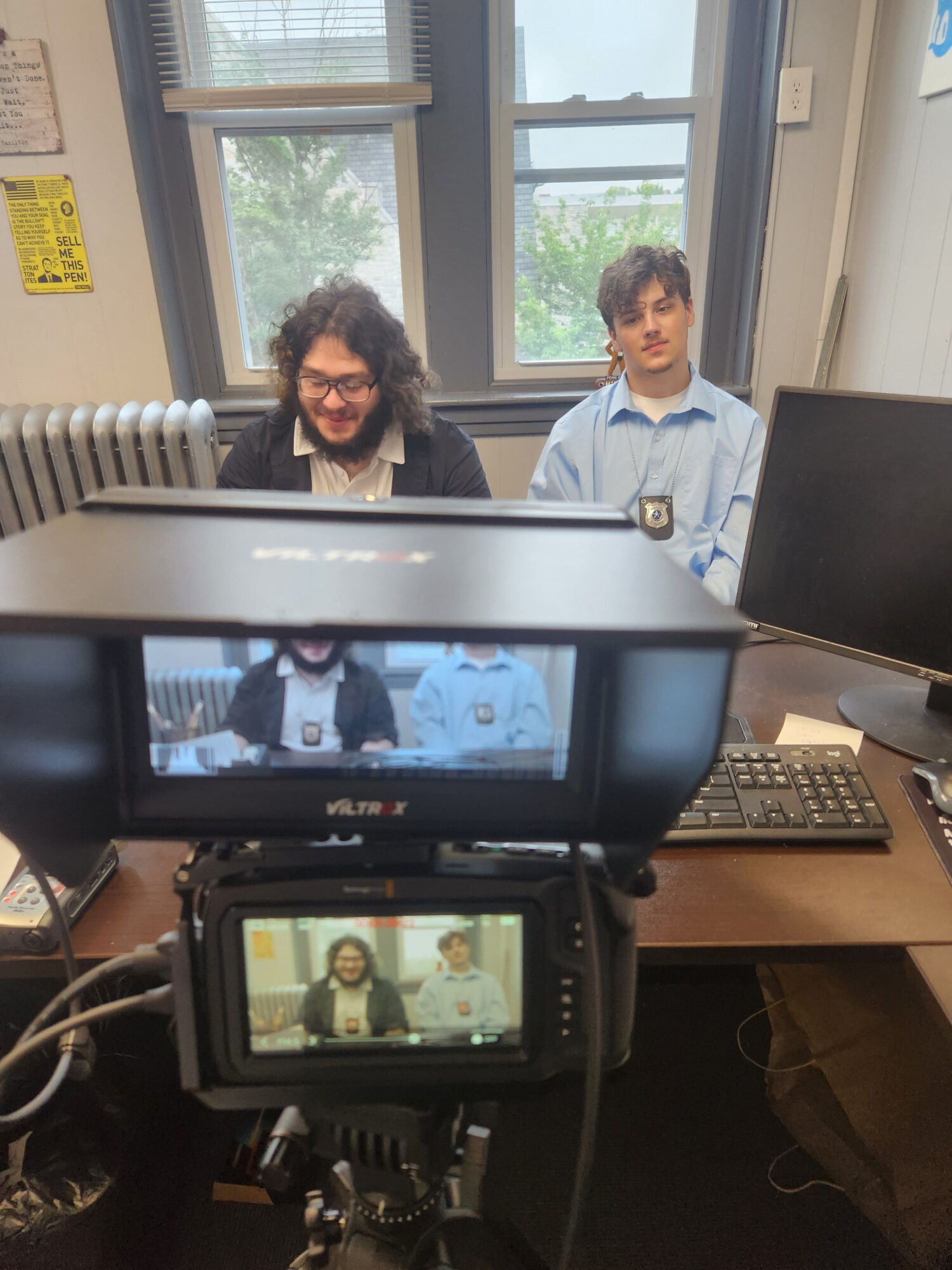 Two people sitting at a desk in an office, with a camera and computer monitor in the foreground, and windows in the background.