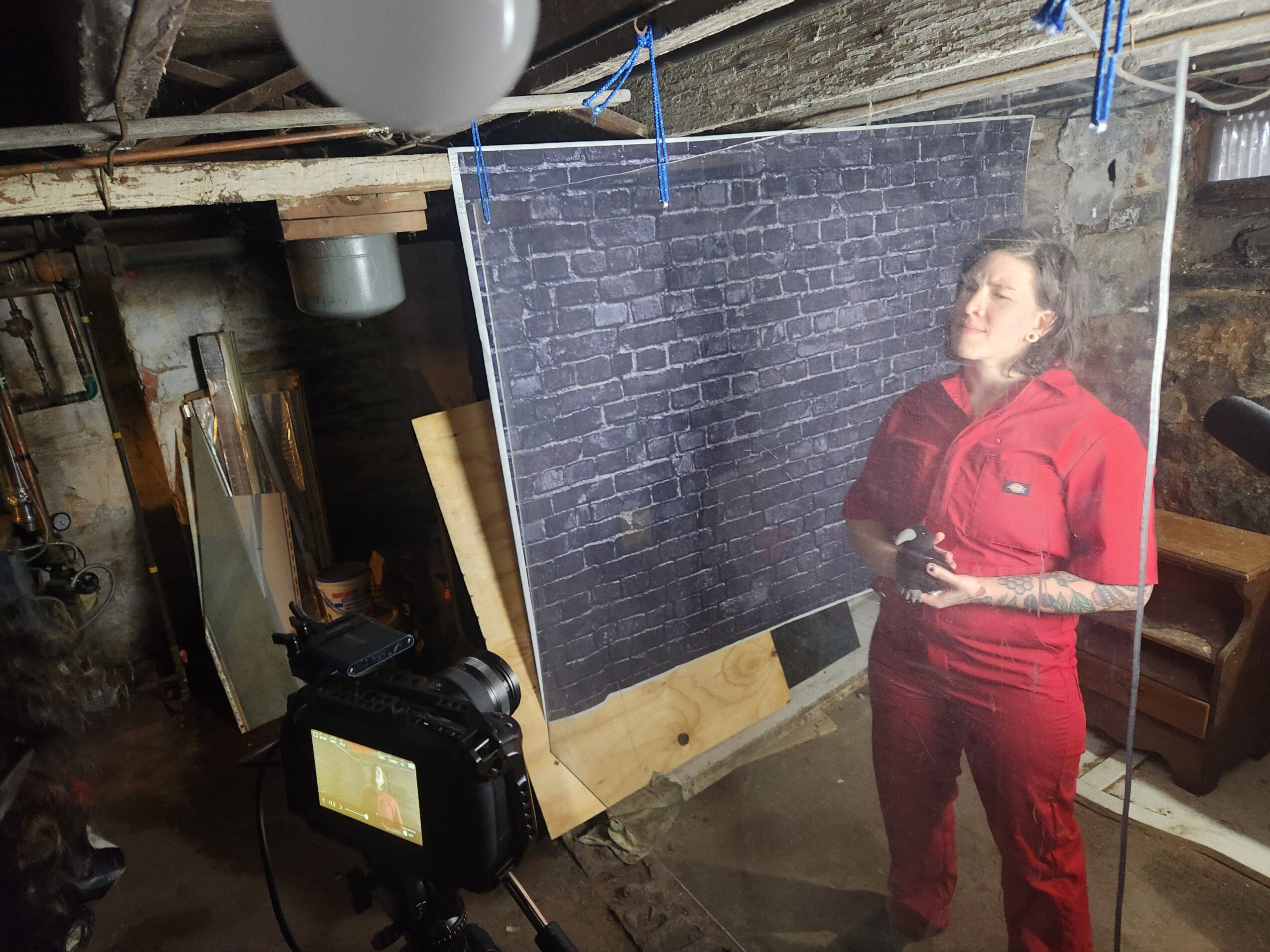 Woman in red jumpsuit holding a tray, standing in front of a black brick wall, in a room with various objects.