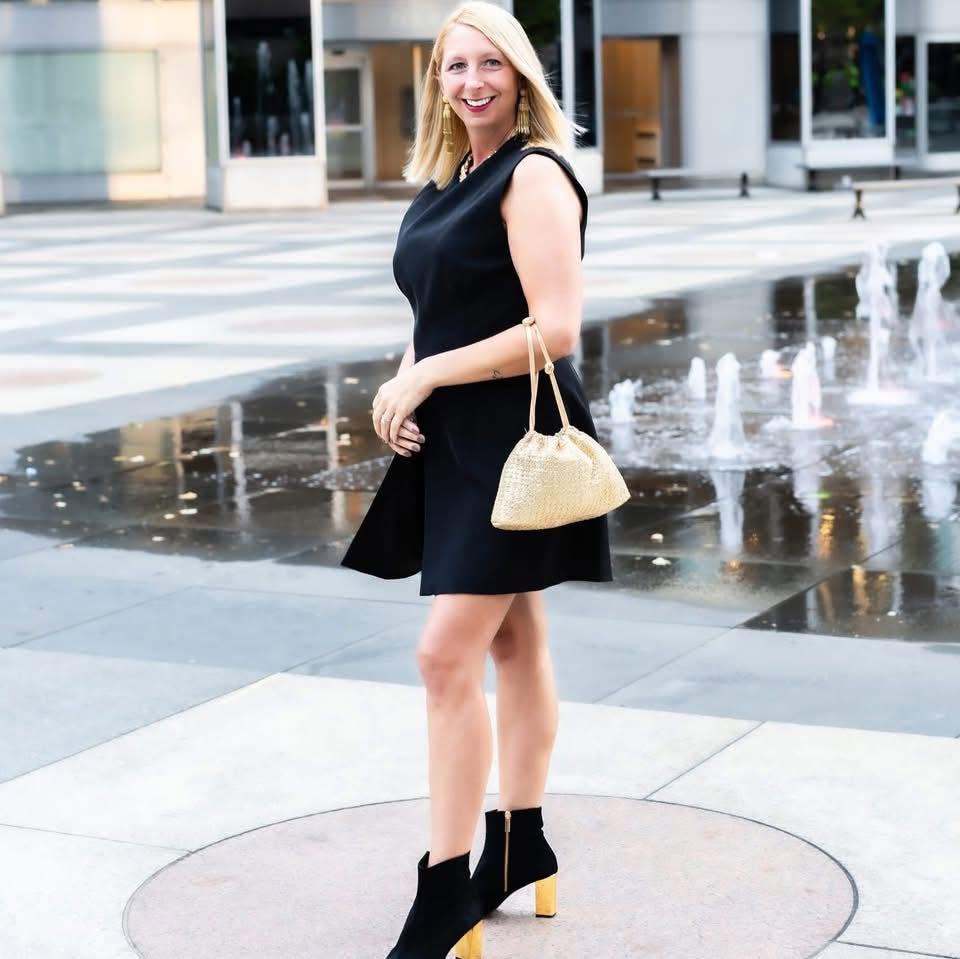 Woman in black dress holding a beige purse standing near water fountain in city street.