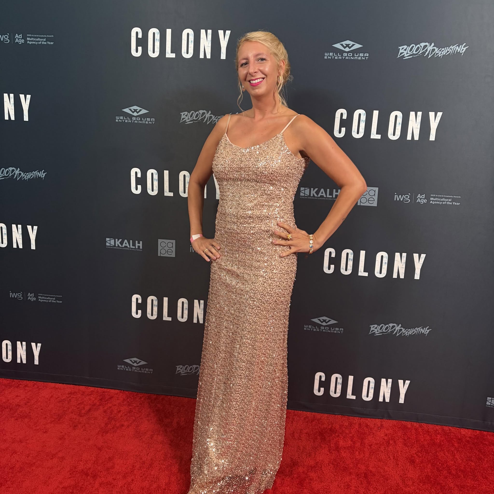 Woman in a sparkly gown standing on red carpet with a dark backdrop reading 'COLONY' behind her.