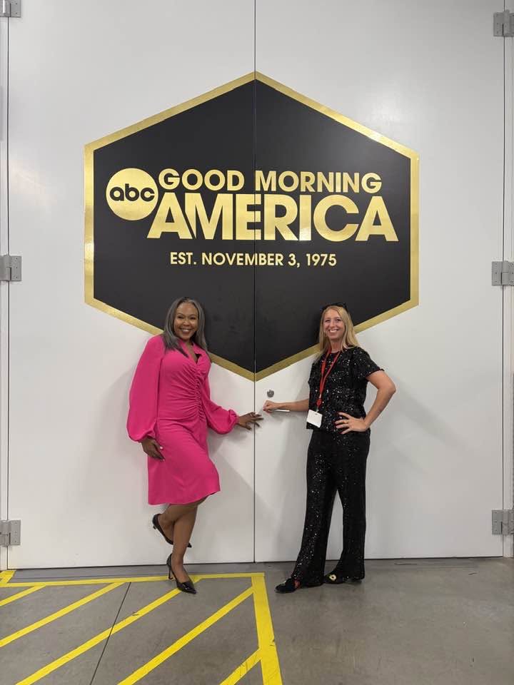 Two women stand in front of a large black and gold sign for ABC Good Morning America, holding hands, smiling.