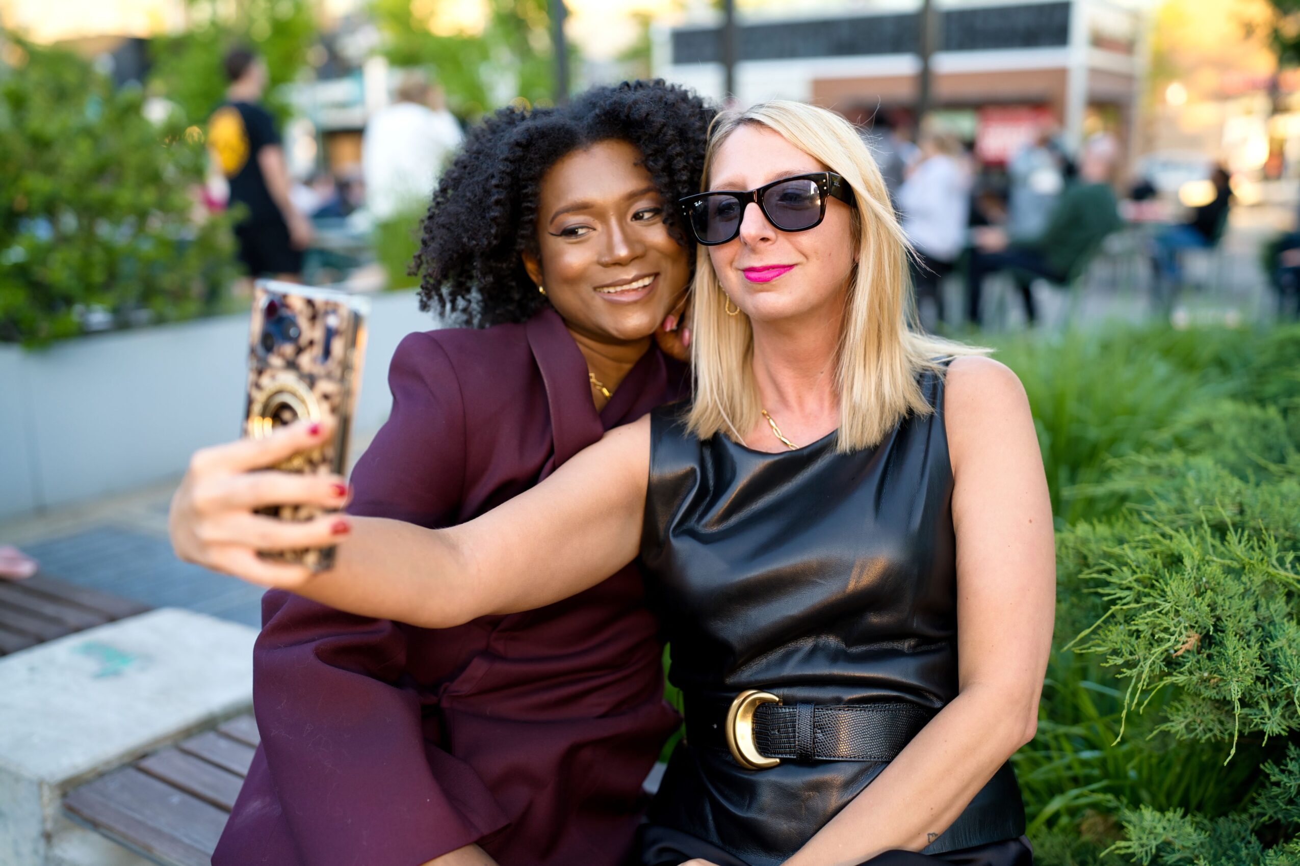 Two women taking a selfie outdoors, one with curly hair and the other with straight blonde hair wearing sunglasses, smiling.