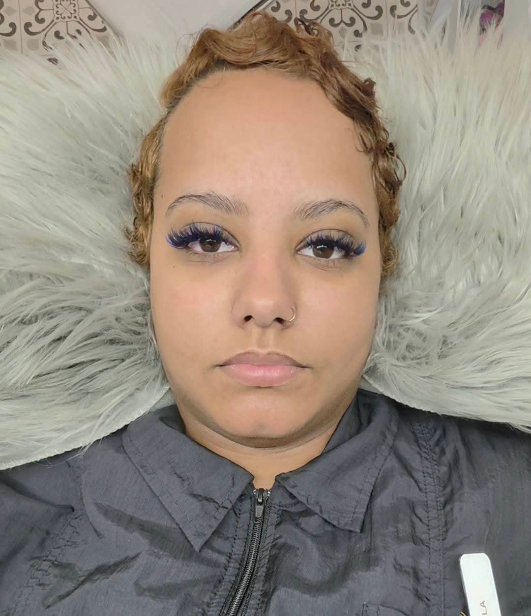 Person with short, curly hair lying on a fluffy pillow, wearing a dark zip-up top, looking directly at the camera.