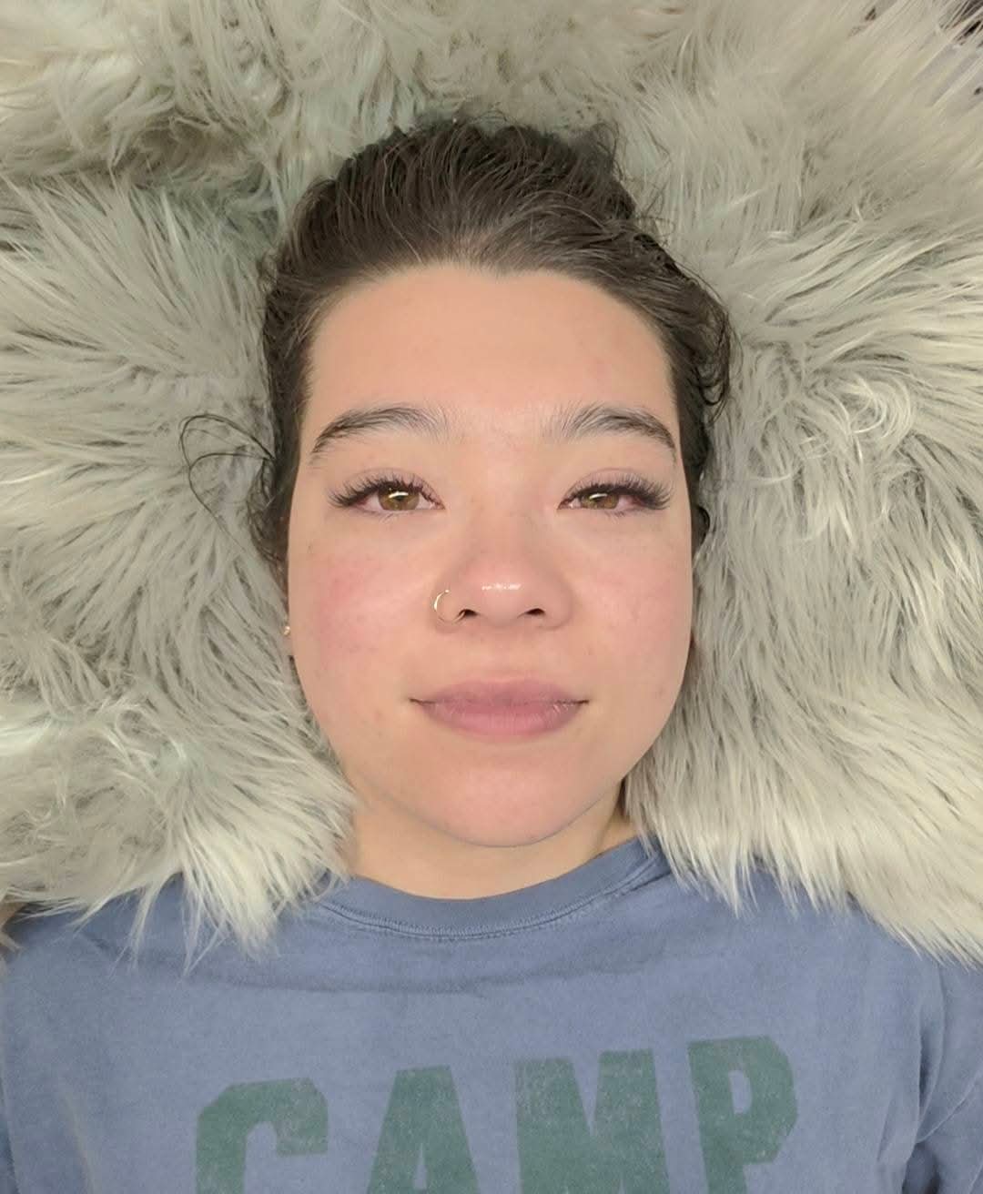 Young woman with dark hair lying on a fluffy white surface, wearing a blue shirt, looking at the camera with a slight smile.