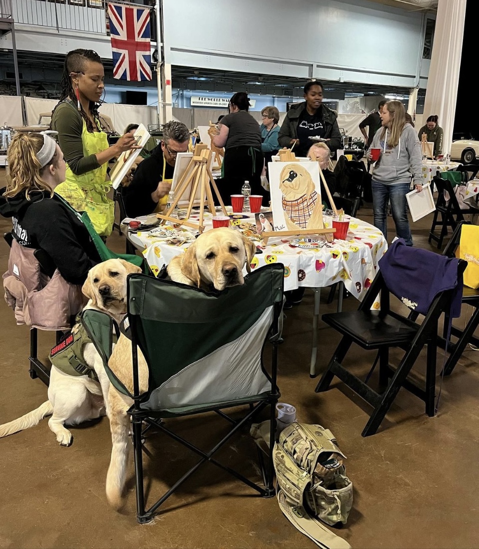 People gather around a table with artwork and supplies, dogs are present, and a British flag is visible in the background.