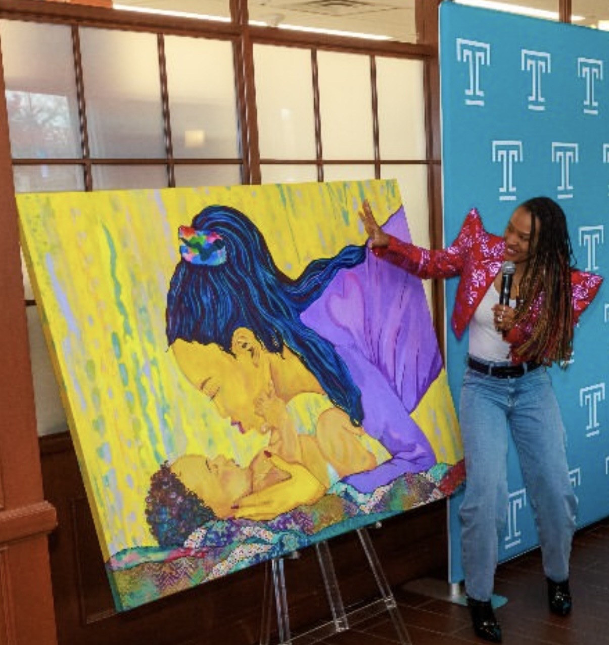 Woman standing next to colorful painting of a woman and child, holding a microphone, in front of a blue background with T logos.