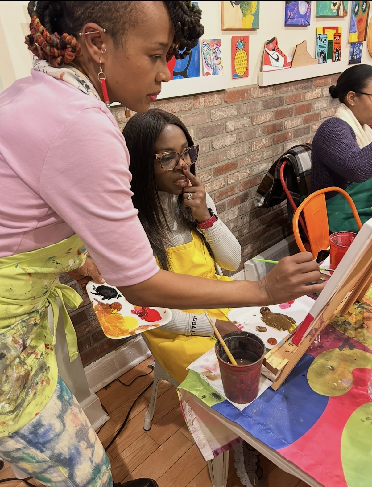 Two women engaged in painting at an art class, with colorful artwork on the brick wall behind them.