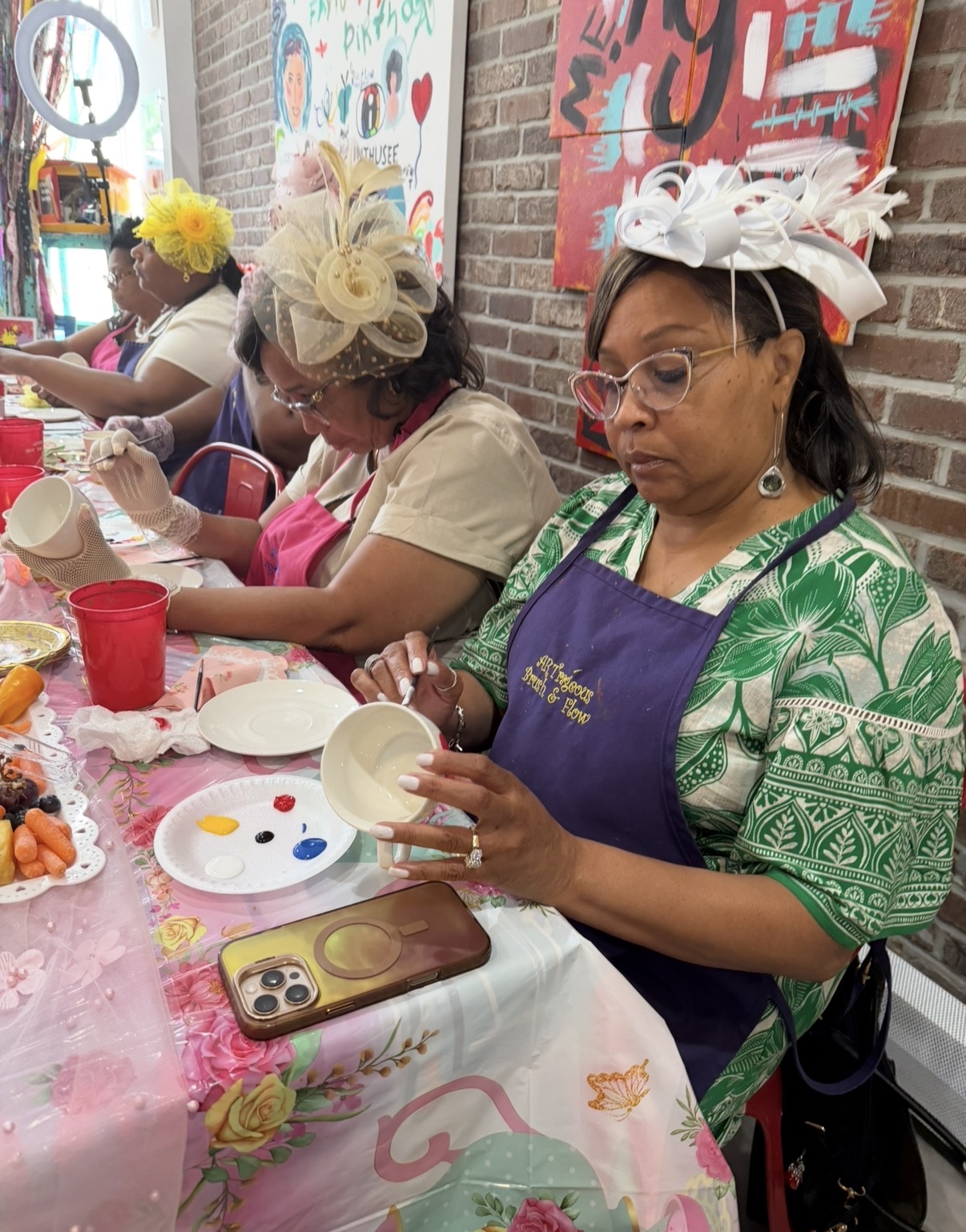 Women wearing decorative hats and aprons sit at a table painting ceramic bowls, with art supplies and food on the table.