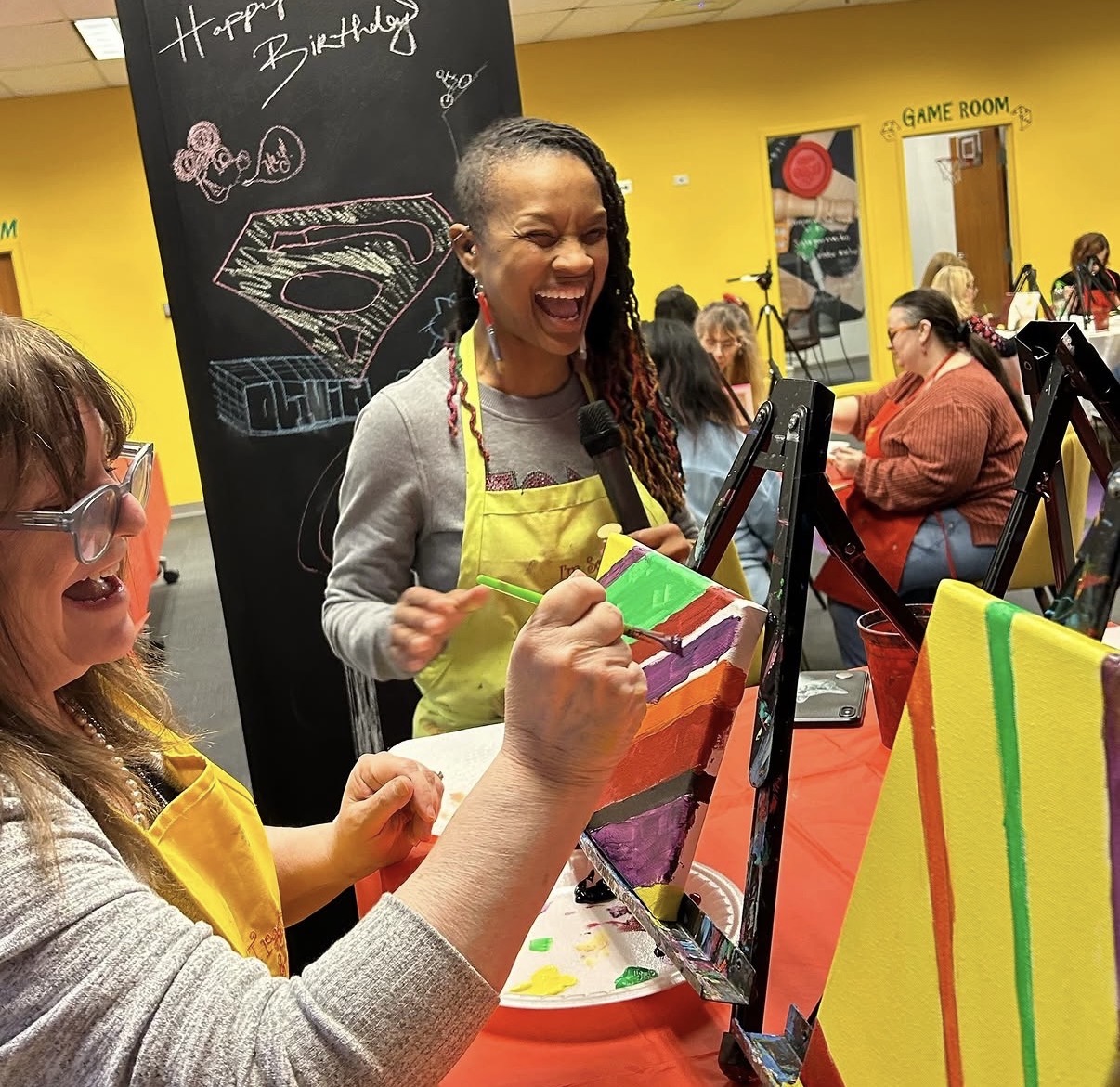 People painting on canvases at a colorful indoor art studio, smiling and enjoying the activity.
