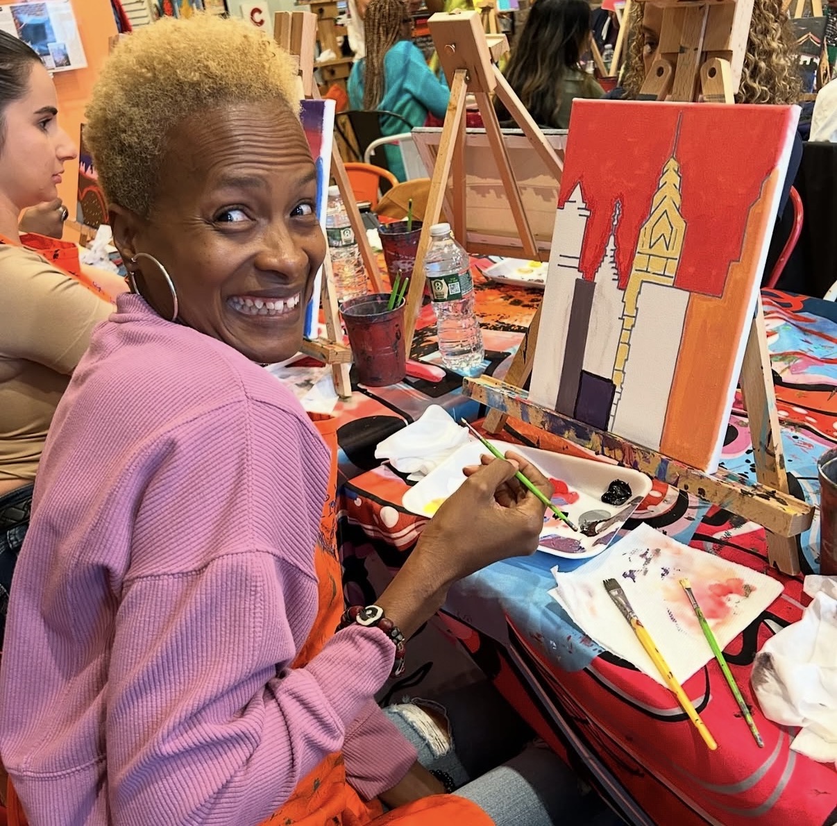 Woman with short blonde hair painting a cityscape on canvas at an art class, smiling at the camera.