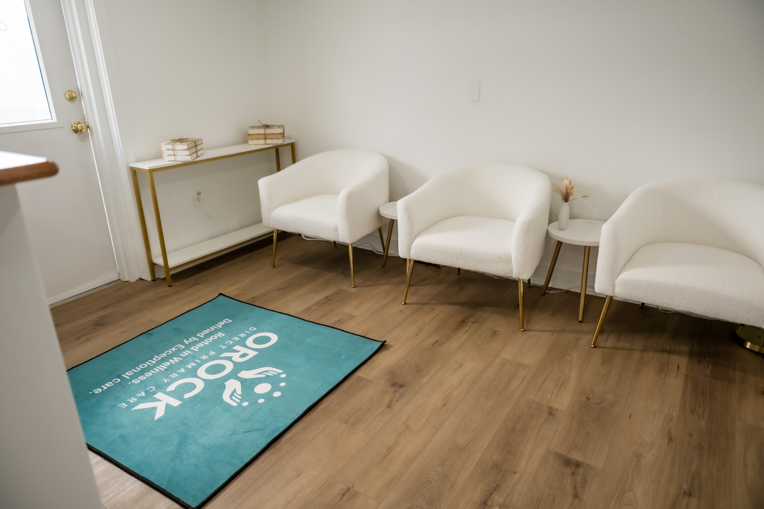 Waiting room with three white chairs, a small table, and a blue rug on wooden floor.