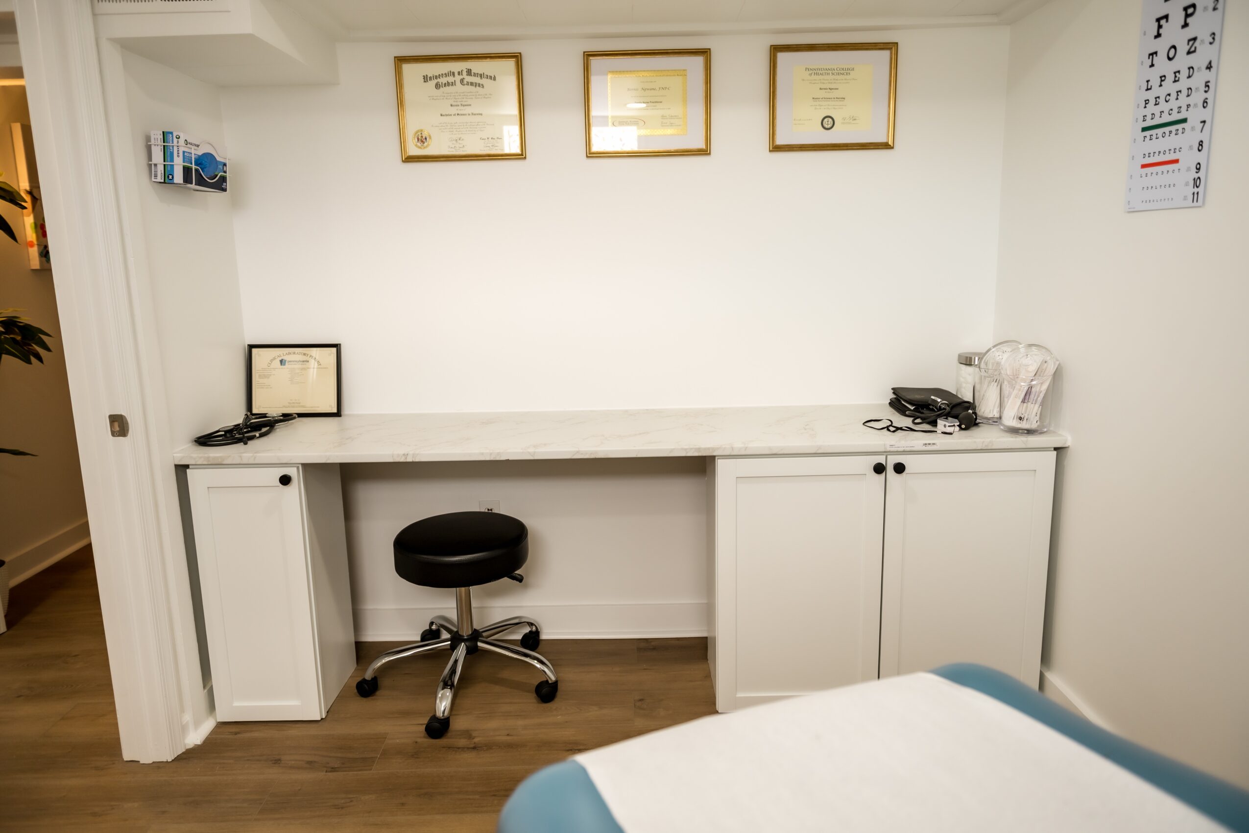 Medical office with a desk, black stool, certificates on the wall, and a bed partially visible in the foreground.