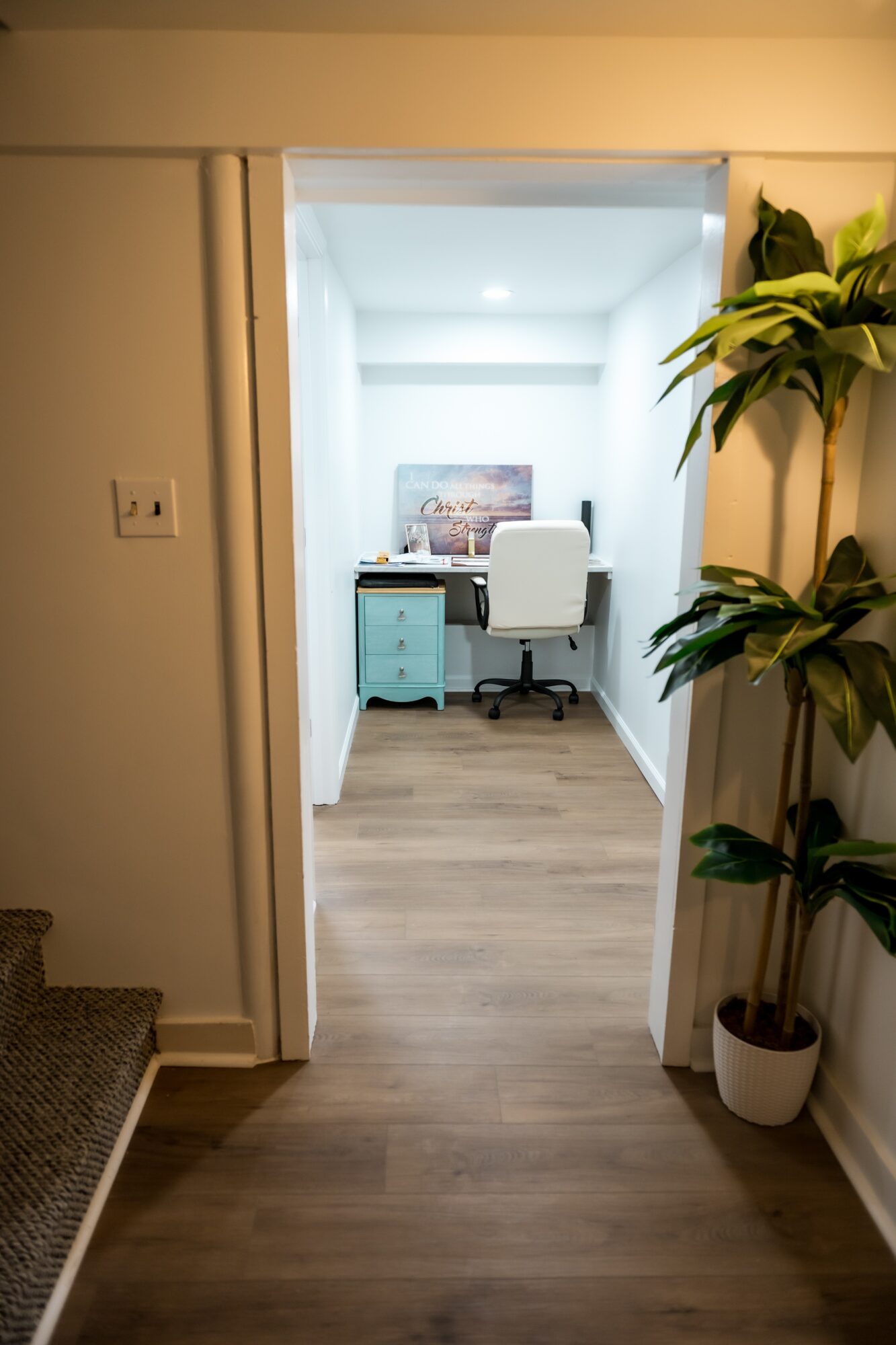 View into a home office with a desk, chair, and a painting on the wall, seen through a doorway with a potted plant nearby.
