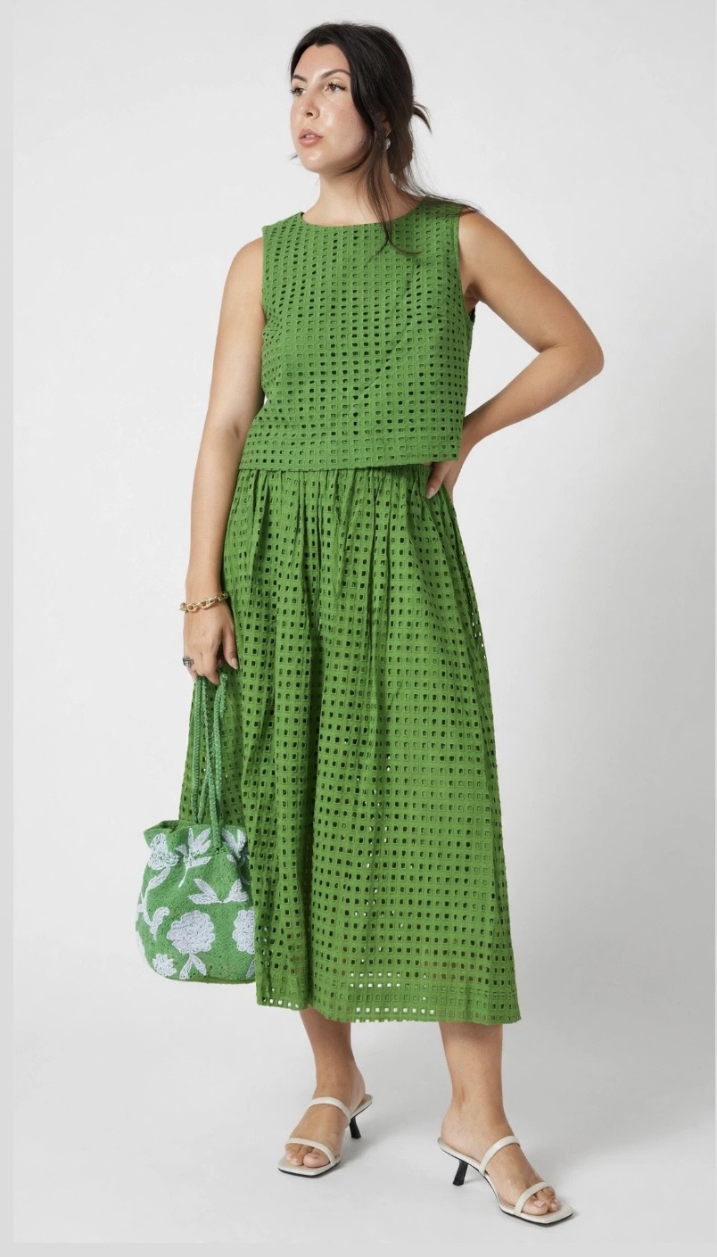 Woman in green polka dot dress holding a patterned bag, standing with one hand on her hip, facing slightly right.