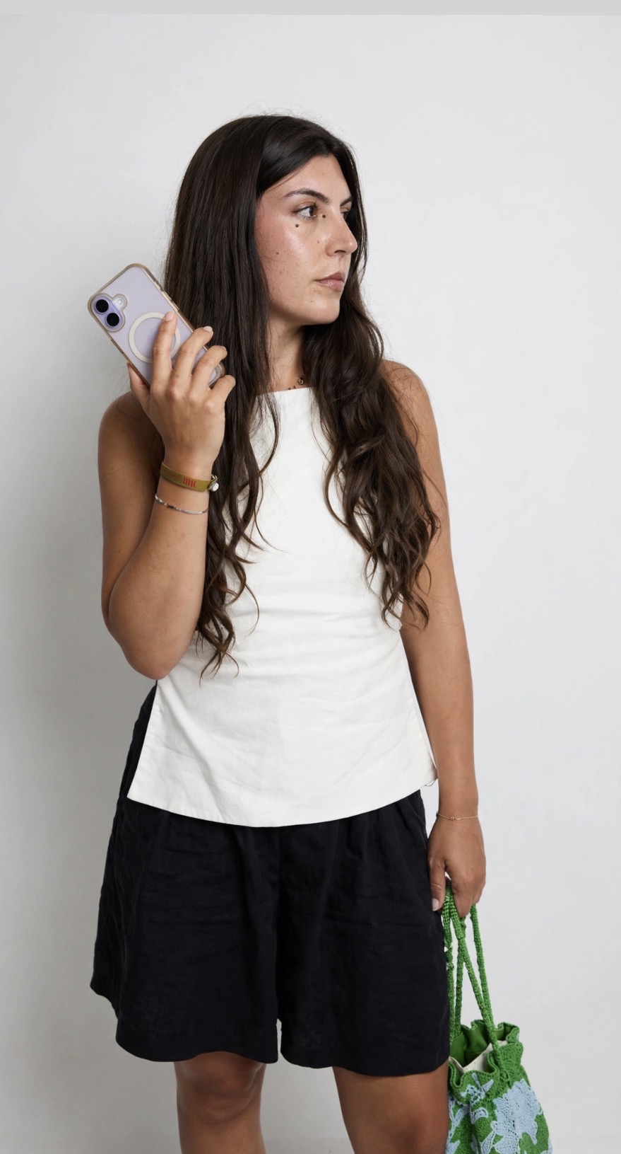 Woman with long dark hair holding a smartphone, wearing a white sleeveless top and black shorts, standing against a plain background.